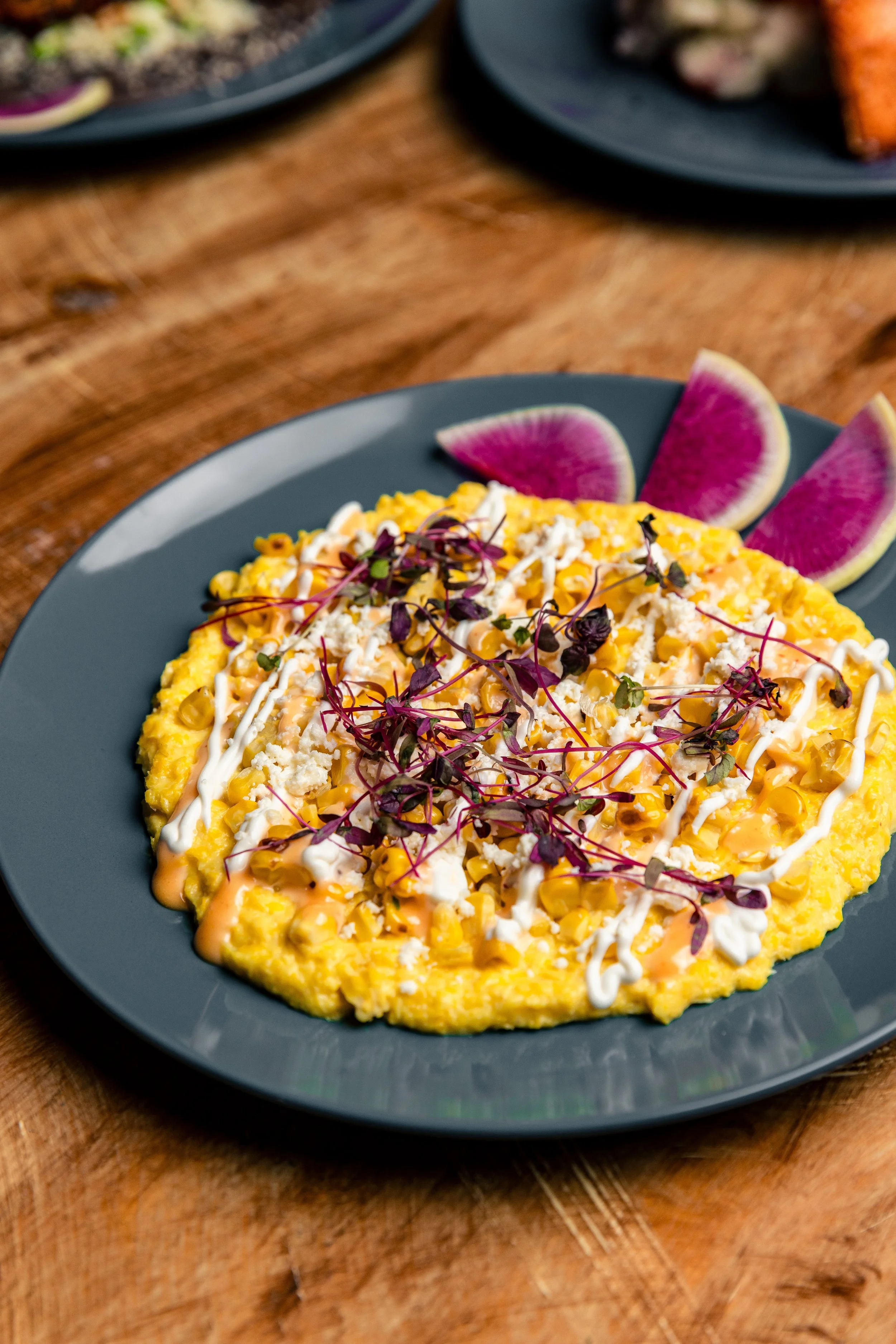 A dish of scrambled eggs topped with microgreens, shredded cheese, and drizzled with sauce and melted cheese, served with slices of watermelon on a gray plate.