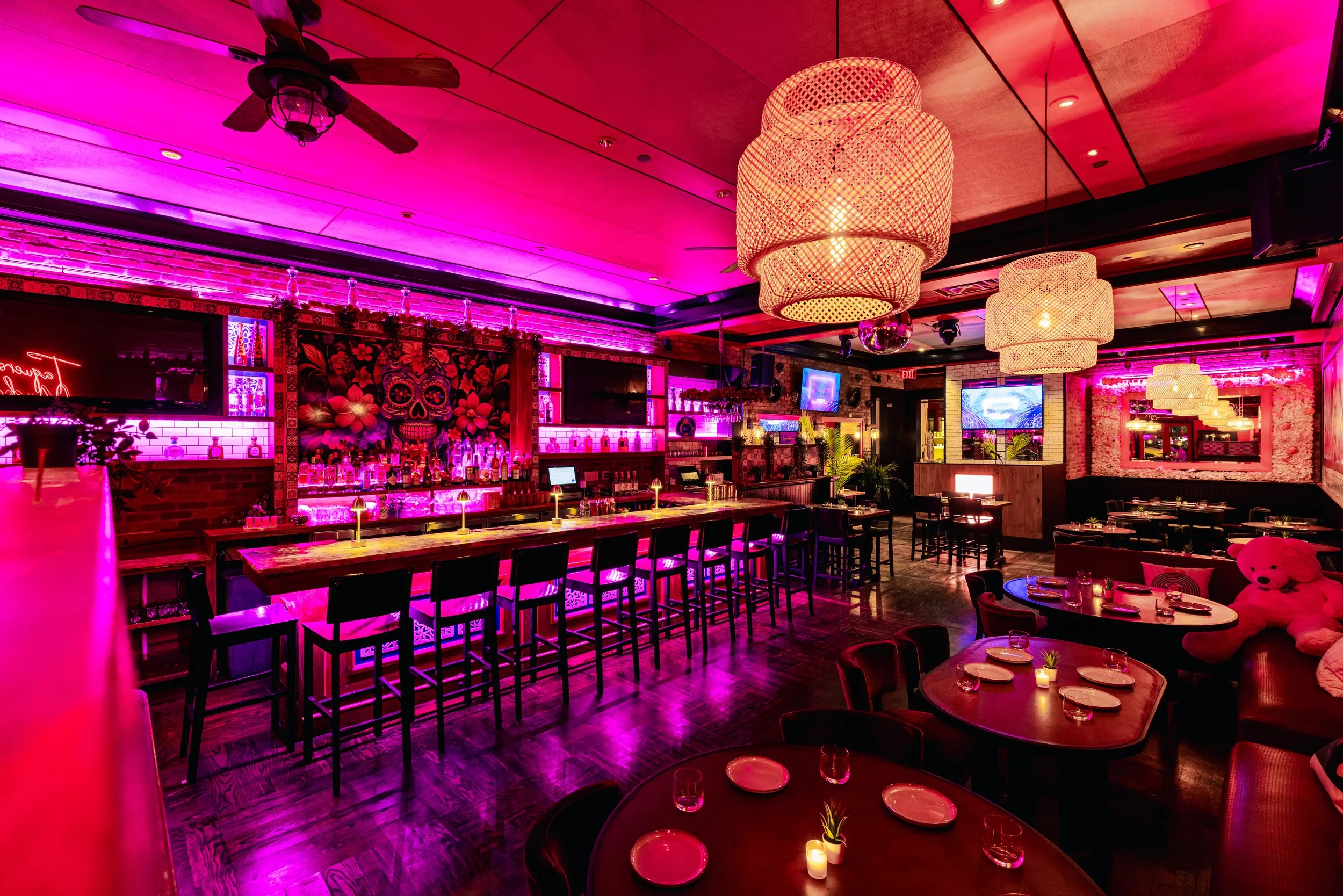 Interior of a vibrant bar or restaurant with pink and purple lighting, large woven hanging lamps, a bar area with bottles and a colorful mural of a sugar skull with flowers, and tables set with candles and glasses.