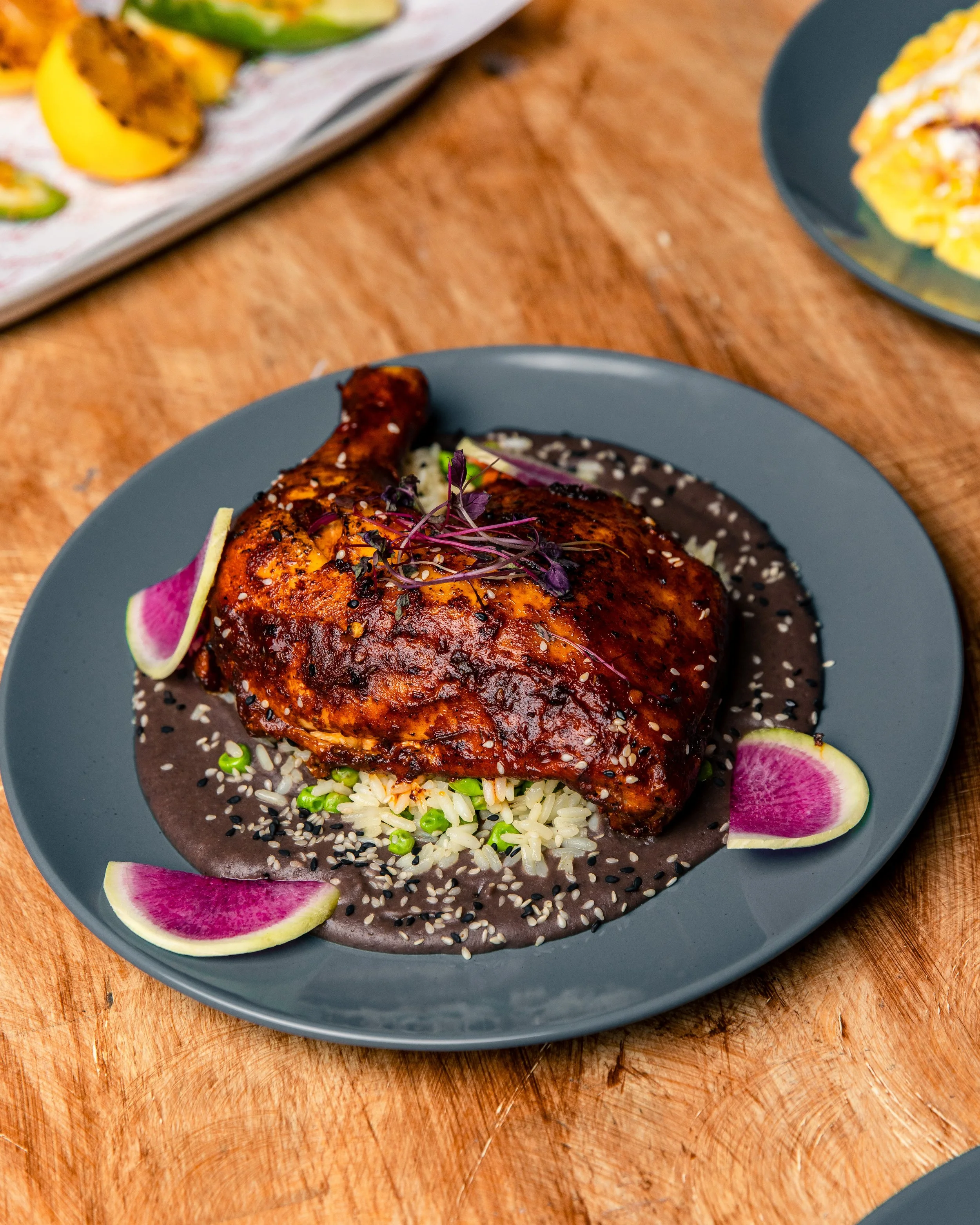 Close-up of a plate with a grilled chicken leg garnished with microgreens, sesame seeds, on a bed of rice with chopped green onions, served with watermelon radish slices and a dark sauce.