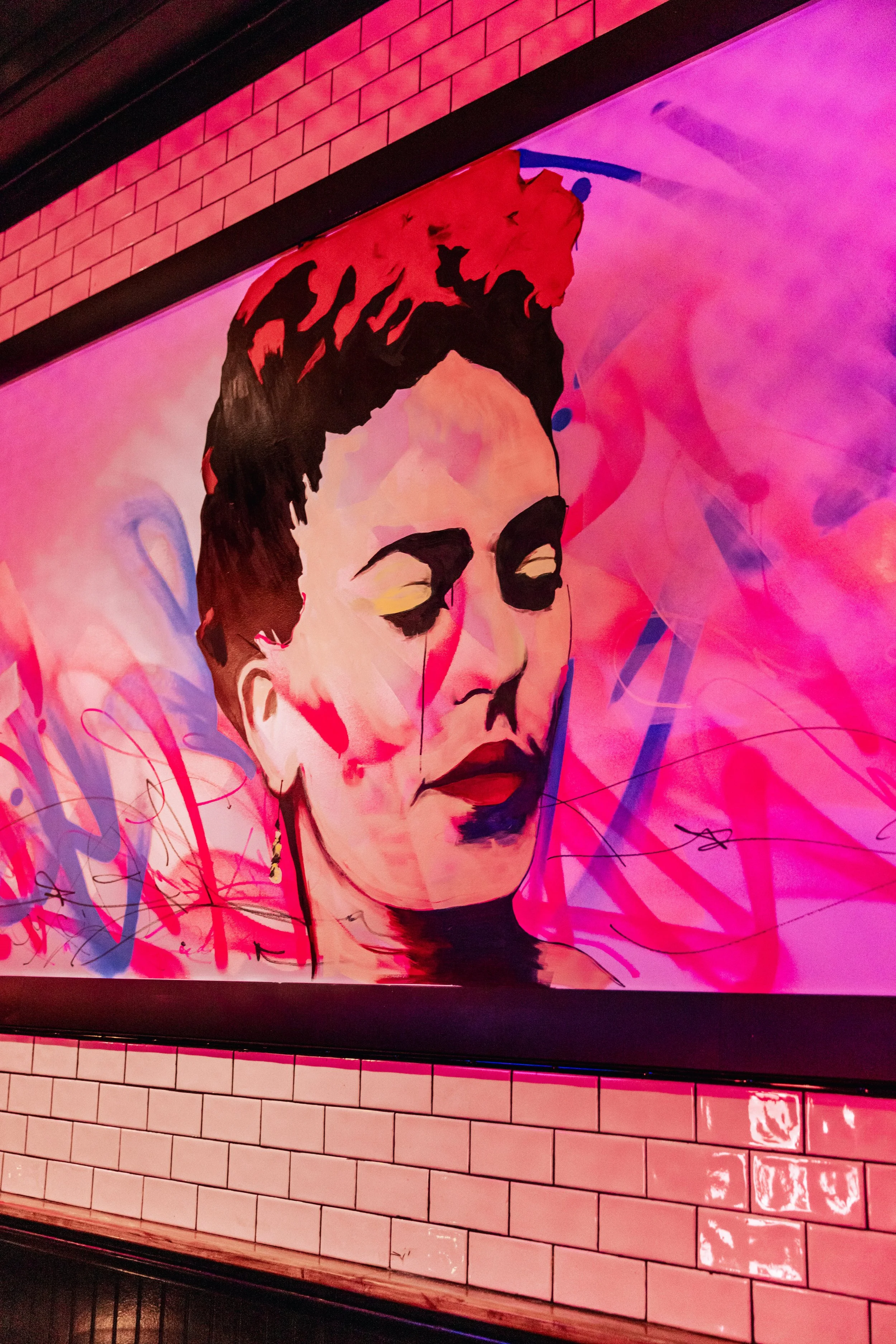 A vibrant, stylized portrait of a woman with closed eyes, dark hair, and bold makeup, painted in bright, neon-like colors with pink, purple, and black tones, displayed on a brightly lit screen against a brick wall background.