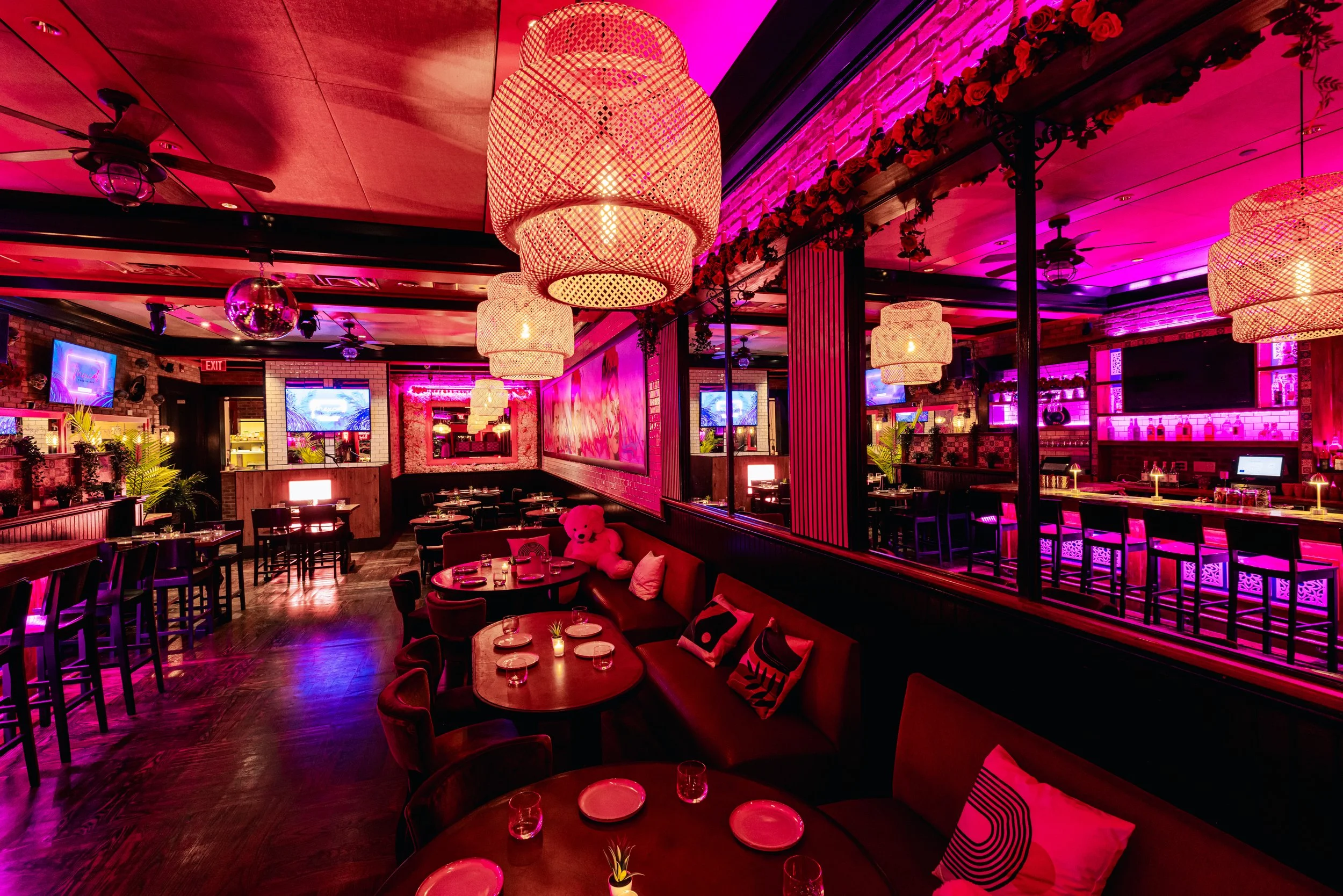 Interior of a restaurant with pink and purple lighting, large woven pendant lamps, a bar area with shelves of bottles, round tables with chairs and candles, and a cozy booth with cushions, pink teddy bear, and small plants.