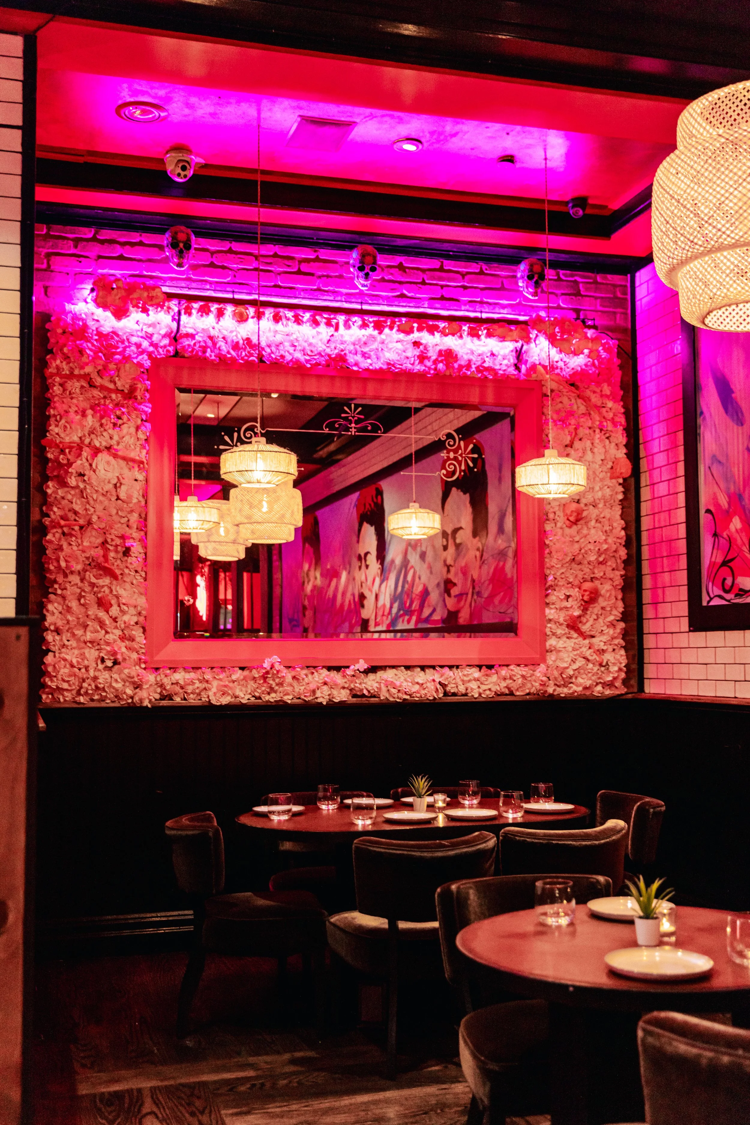 Interior of a restaurant with pink and purple lighting, decorated with flower frame, hanging lamps, and skull decorations on the ceiling. Tables are set with glasses, plates, and small candles.