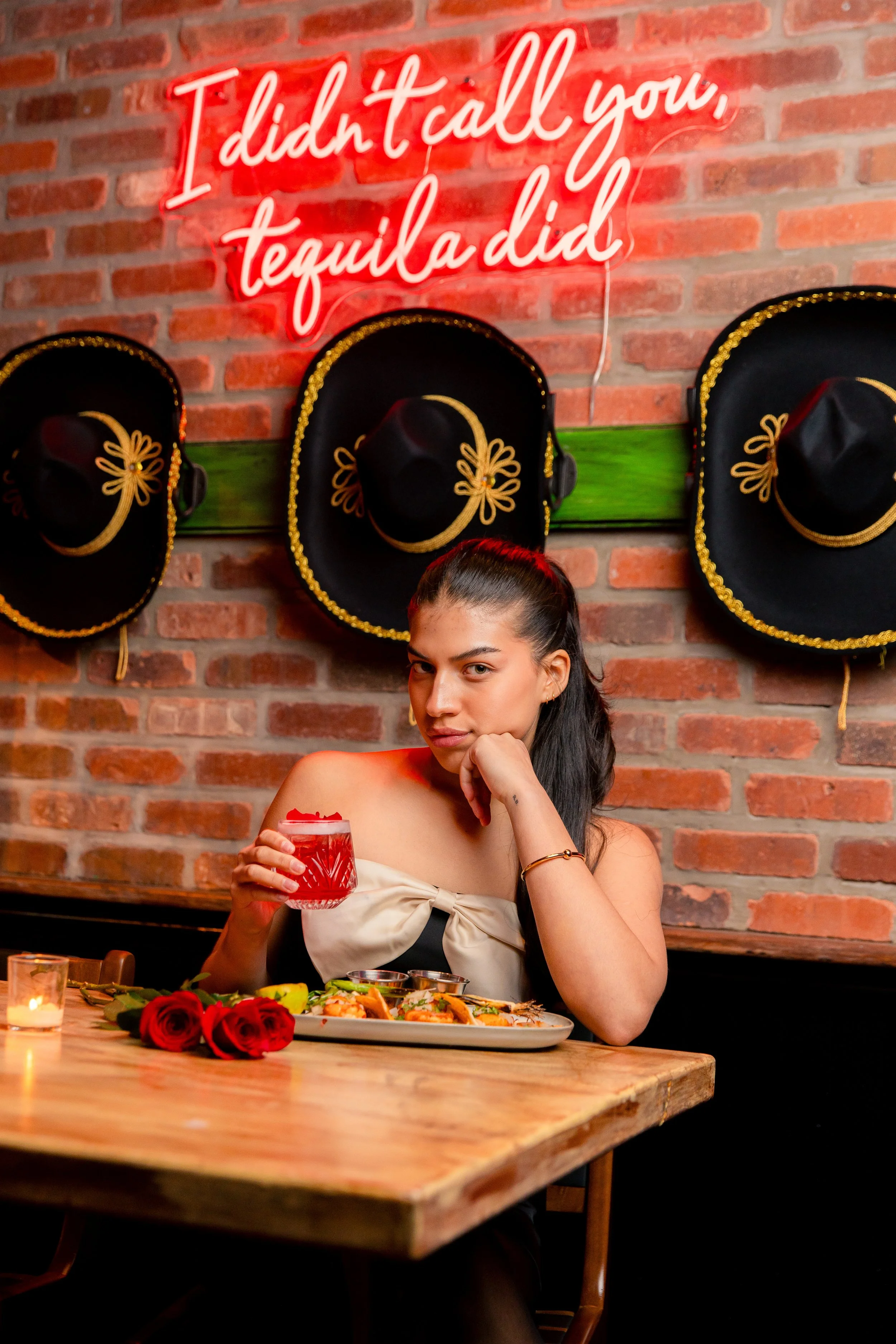 A woman is sitting at a wooden table in a restaurant, holding a glass of red drink with a straw. She has dark hair tied back, is wearing a beige strapless top with a bow detail, and has a gold bracelet on her right wrist. There are red roses and a pl