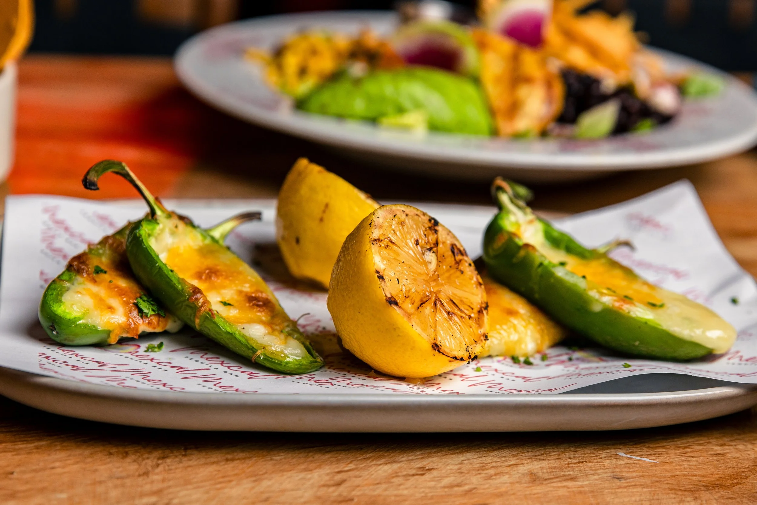 Grilled jalapeño peppers with melted cheese and a grilled lemon wedge on a white paper underlayment, with a plate of salad in the background.