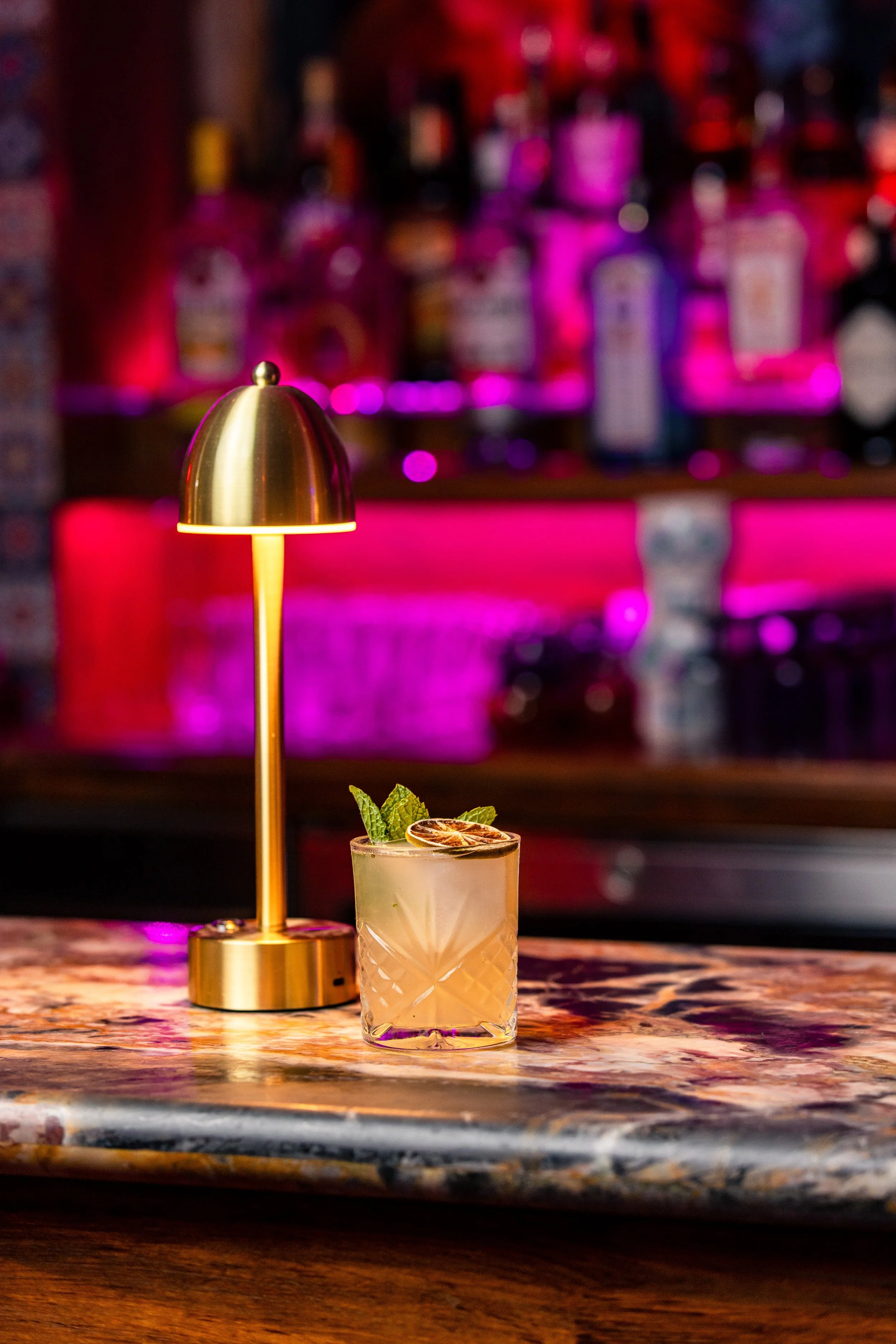 A cocktail drink garnished with mint leaves and dried citrus slice on a patterned glass, placed on a bar counter, with a brass-colored small lamp beside it. The background features a bar with bottles and pinkish-purple lighting.