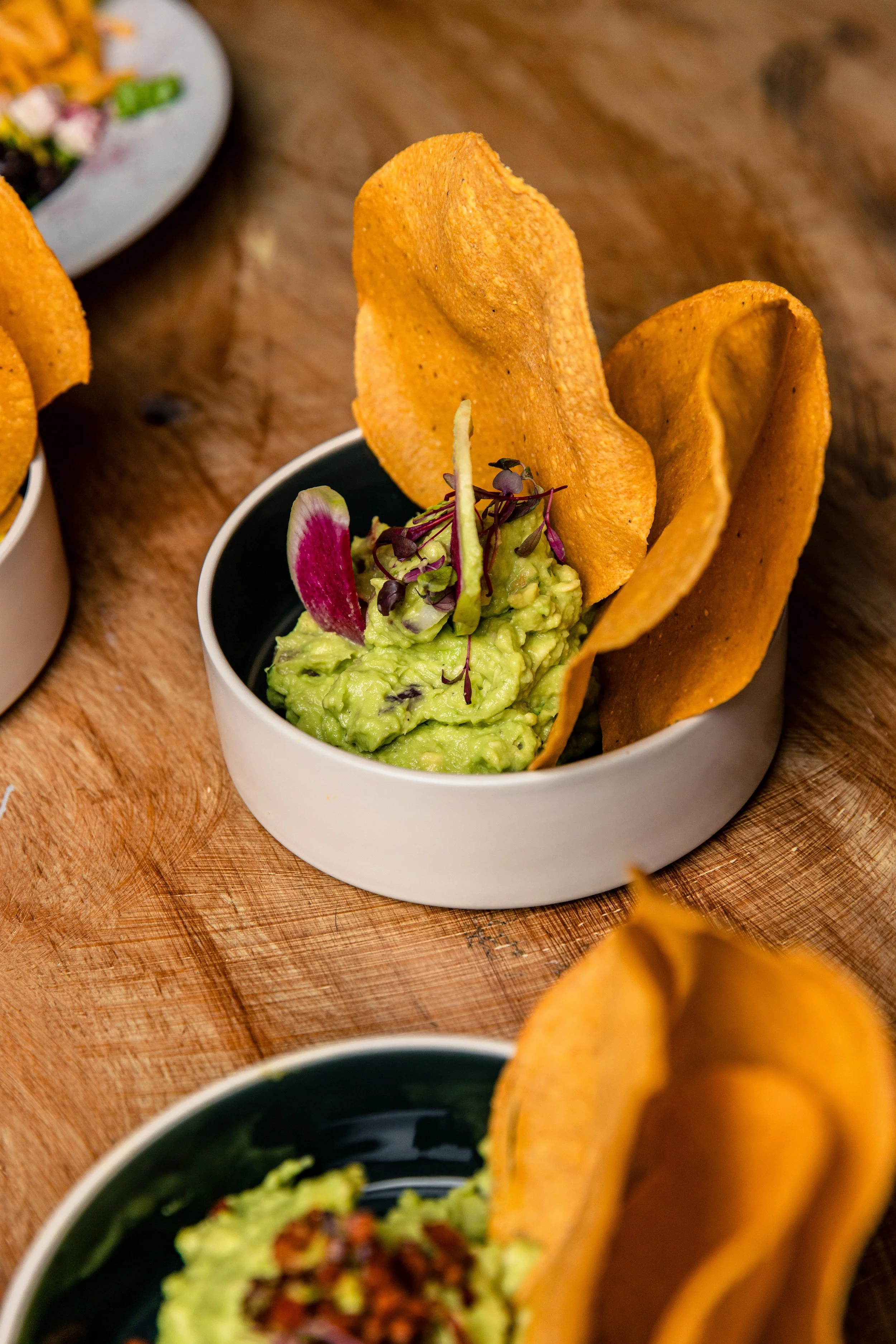 Close-up of a small bowl of guacamole with microgreens and a radish slice, served with two crispy taco shells.