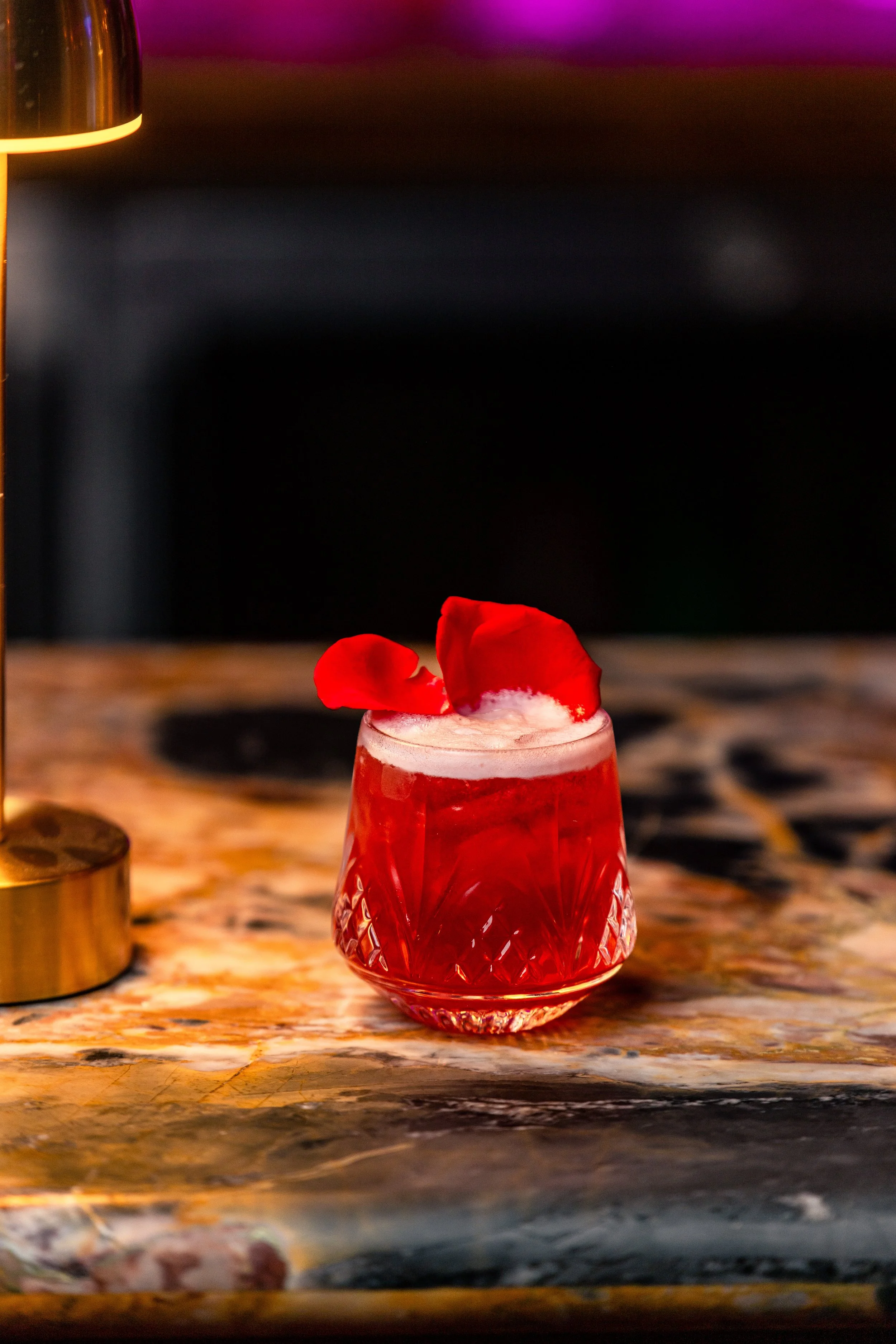 A red cocktail with rose petals garnishing the top, served in a decorative glass on a marble tabletop.
