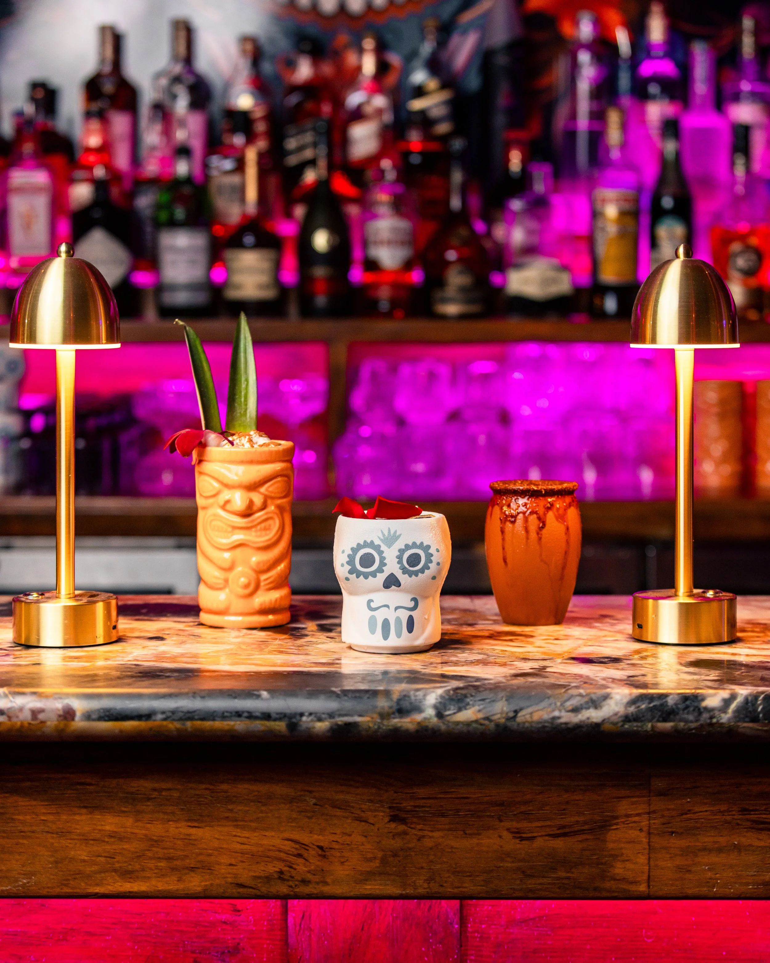 A bar with various bottles of alcohol in the background, illuminated by pink and purple lighting. In the foreground, a tiki glass with a tropical drink, a skull-shaped cup with a skull face design, and another ornate cup are on the bar counter. Two small gold lamps with curved shades flank the drinks.