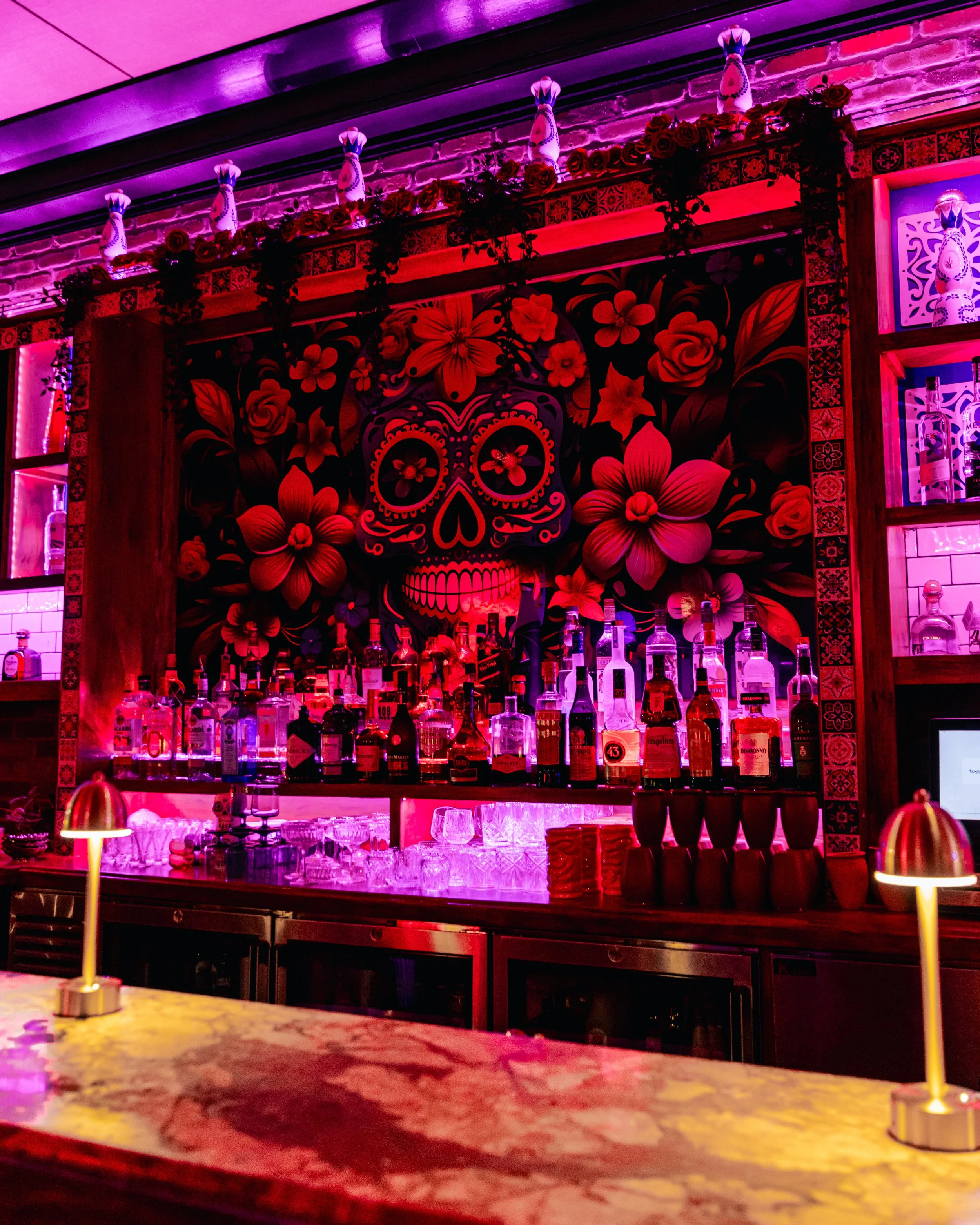 Colorful bar with a decorated skull mural and bottles of alcohol. Pink and purple lighting create a vibrant atmosphere.