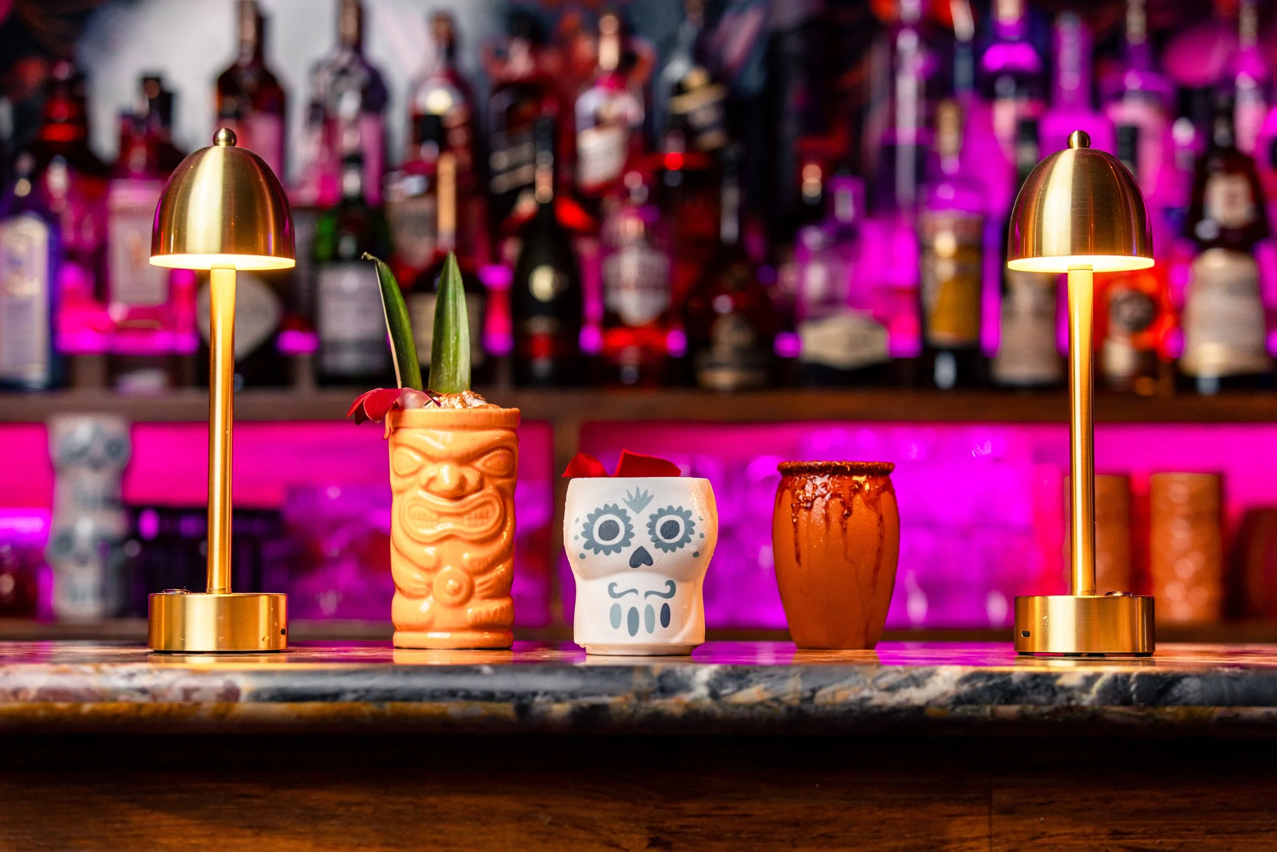 Bar scene with three gold lamps and colorful mugs with Day of the Dead themes, set against a vibrant pink-lit background of liquor bottles.