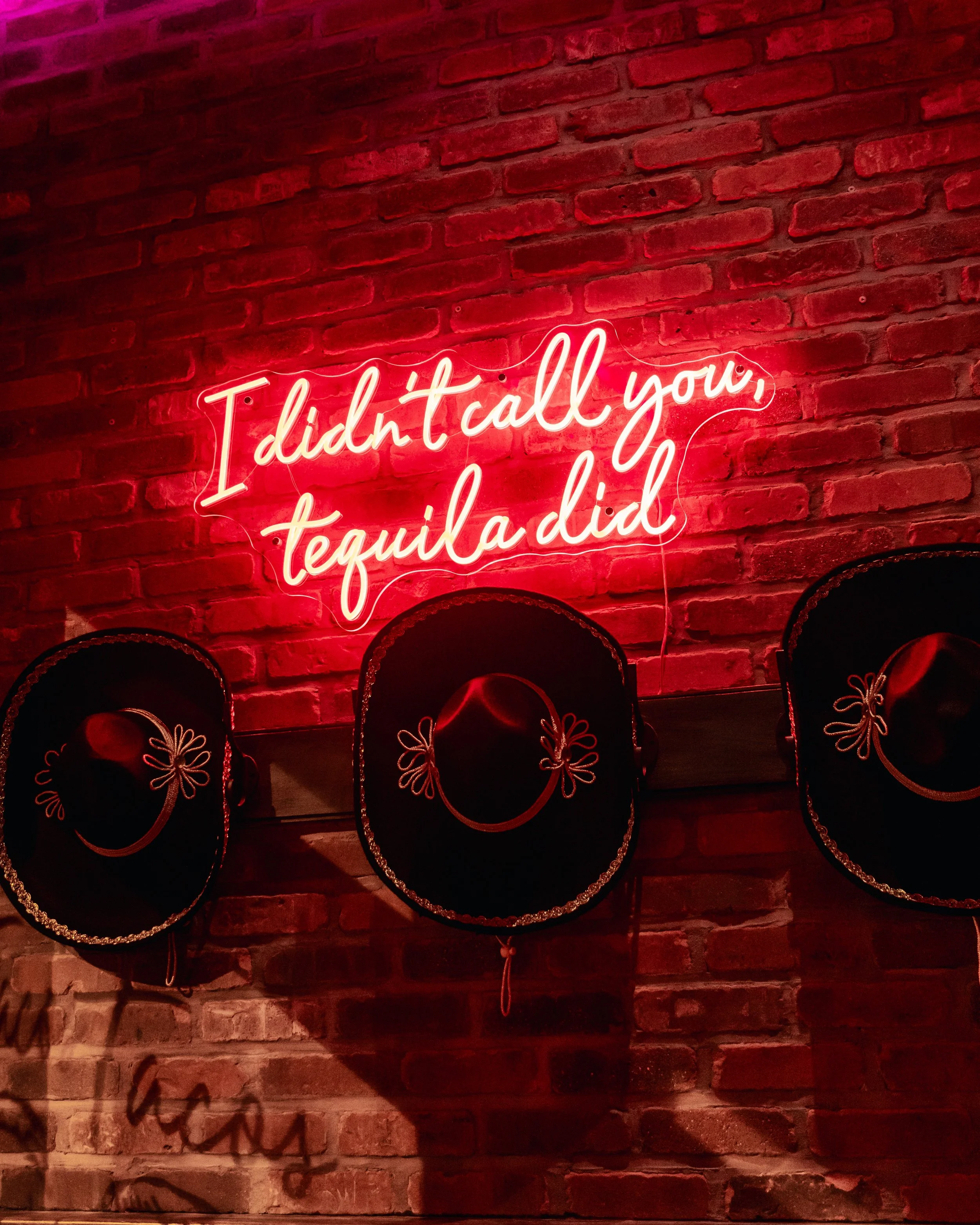 Neon sign on red brick wall displays the phrase 'I didn't call you, tequila did'.