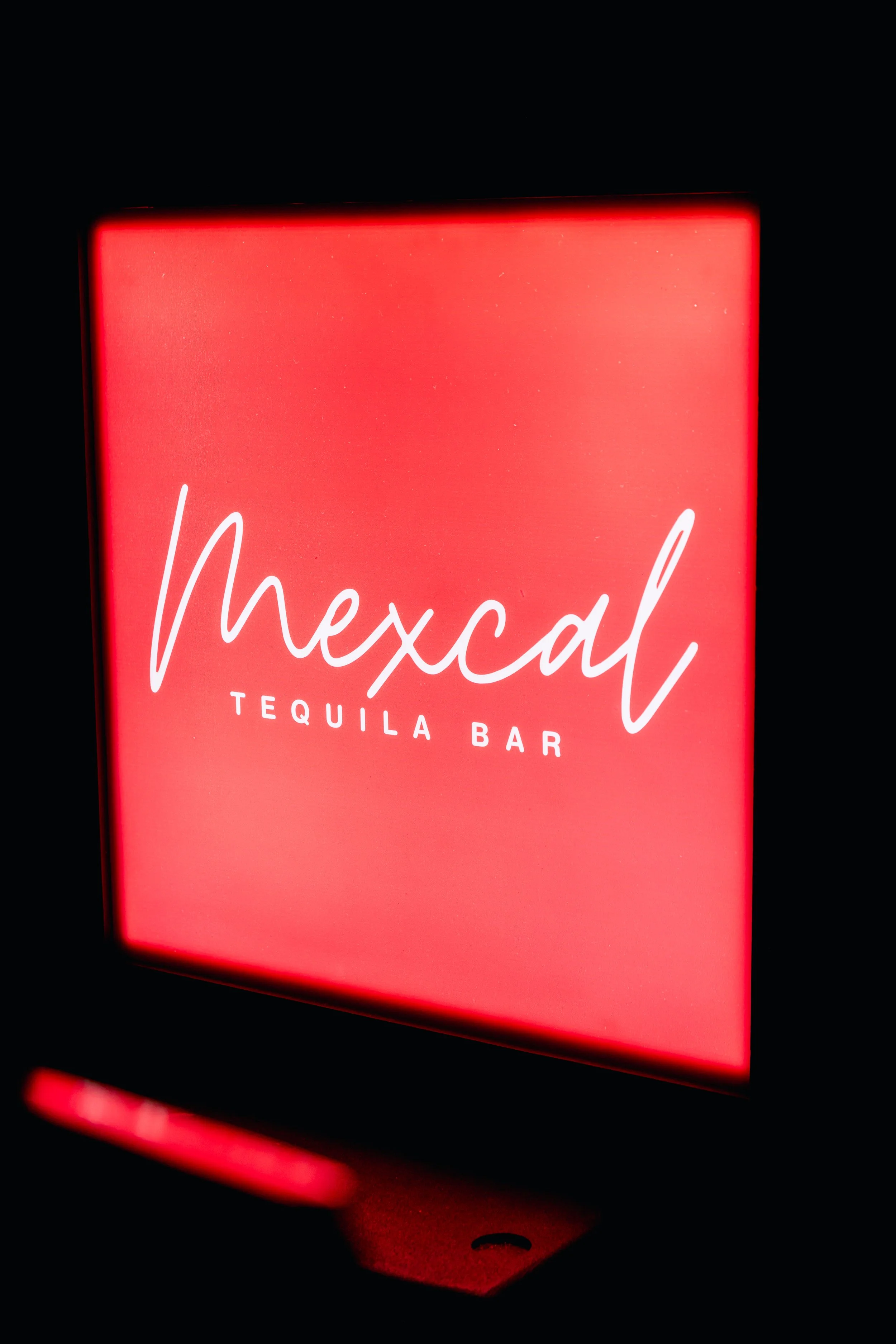 A neon sign with the words "mexcal" and "TEQUILA BAR" in white script, glowing red against a black background.