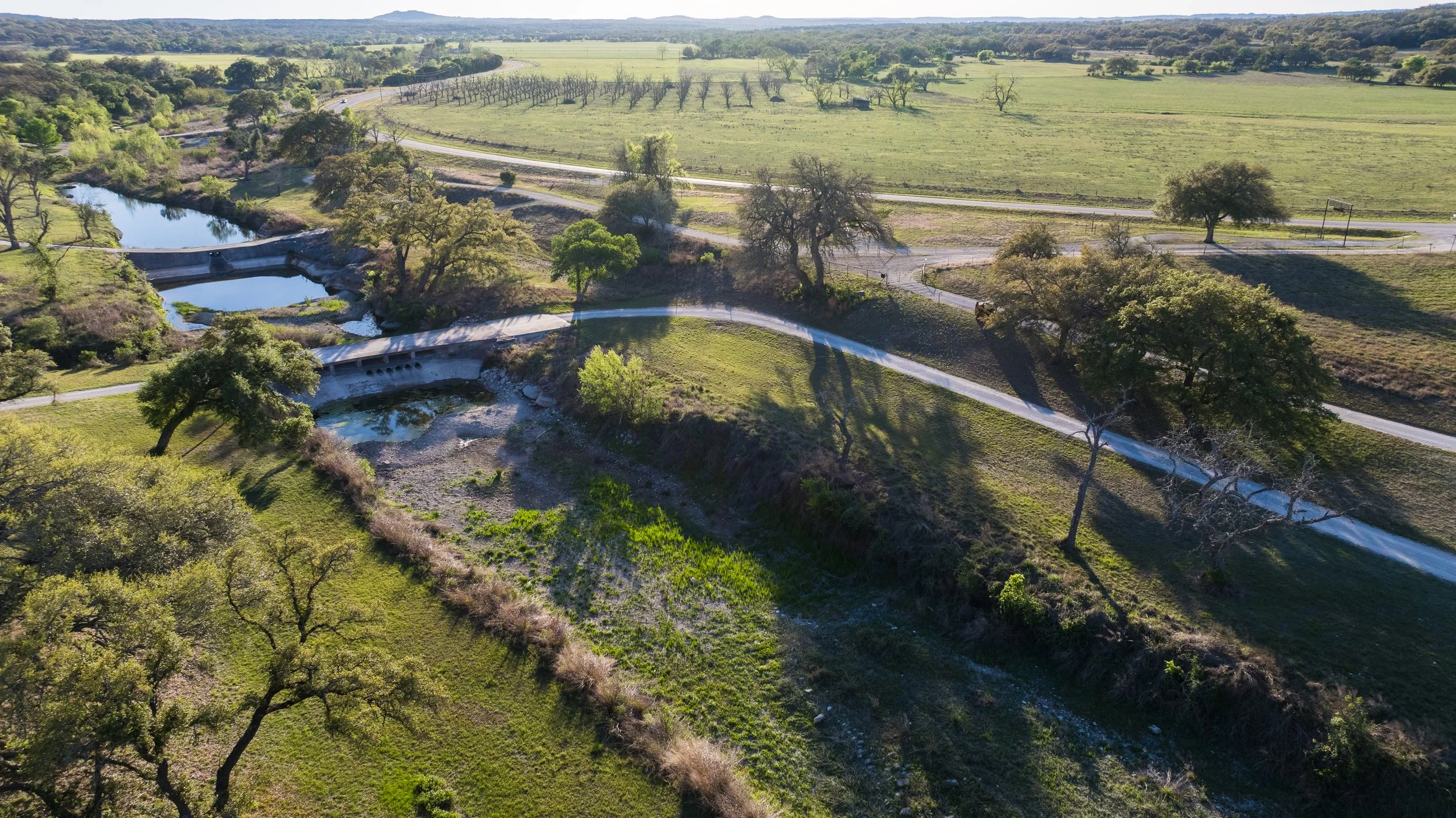 Aerial view of a landscape with a small river or creek, a dam, winding roads, trees, open grassy fields, and distant hills under a clear sky.