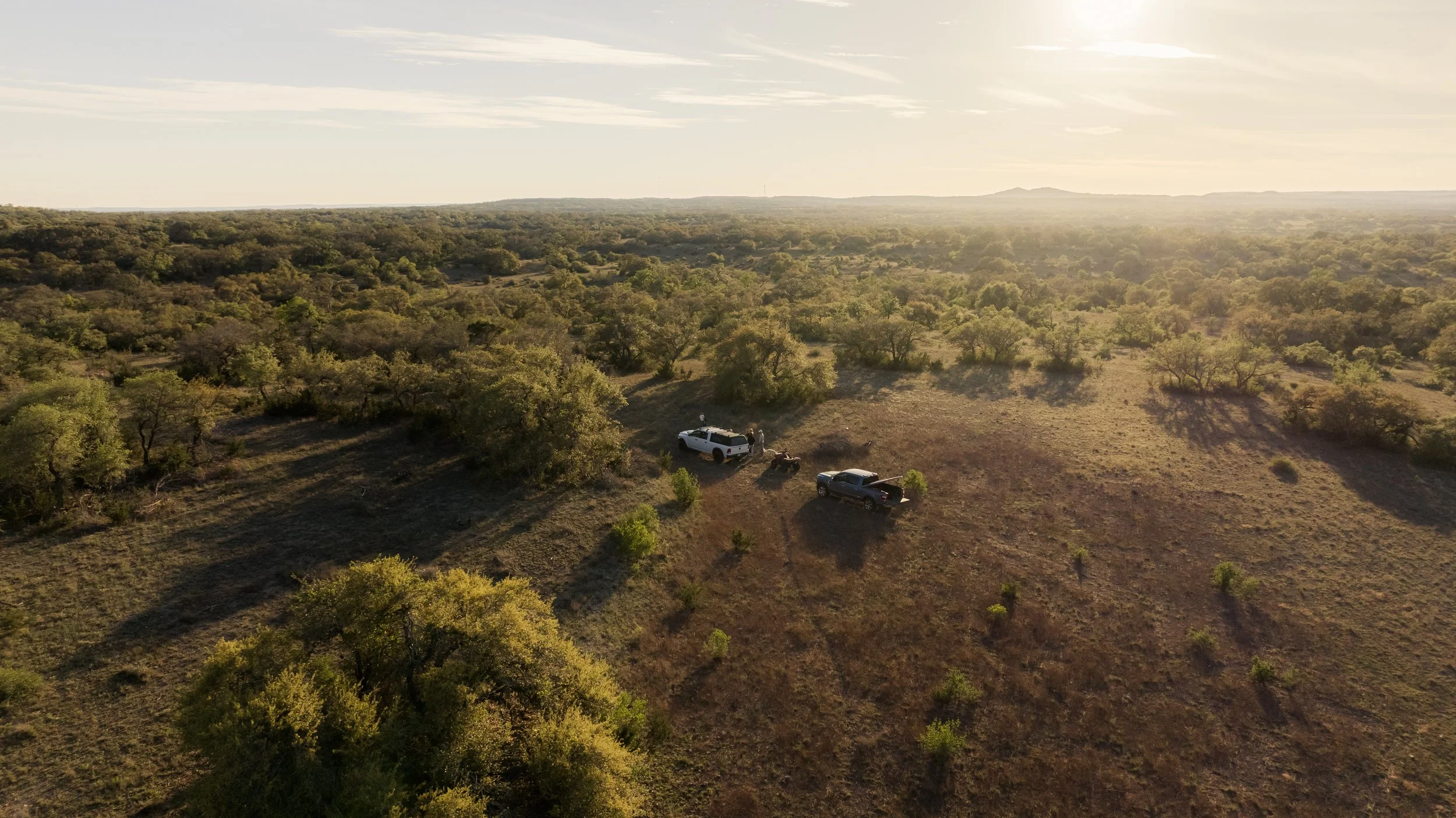 Aerial view of a rural landscape with trees, a patch of open land, and three pickup trucks parked near some bushes during sunset.