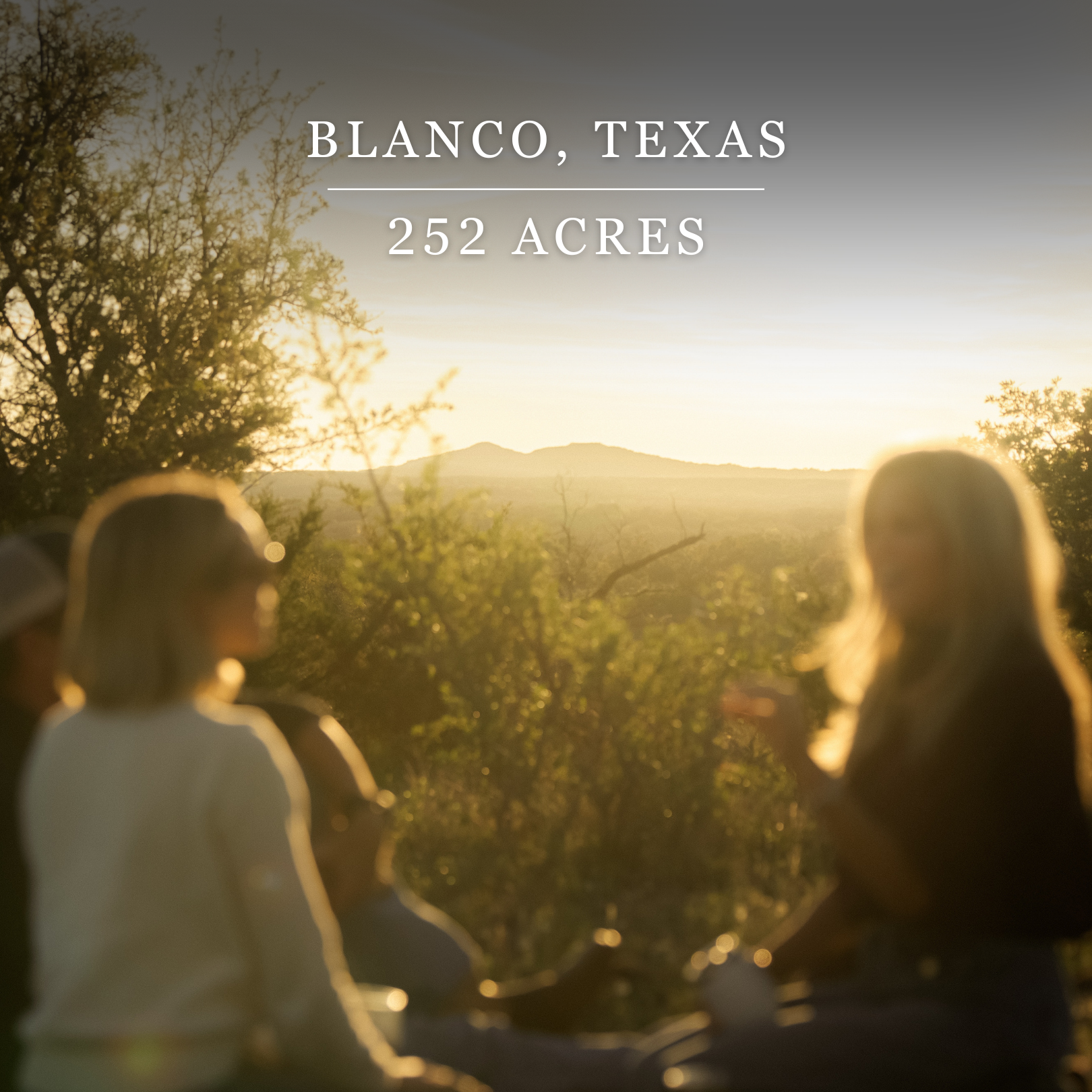 People gather outdoors at sunset in a scenic landscape with hills and trees in Blanco, Texas, covering 252 acres.