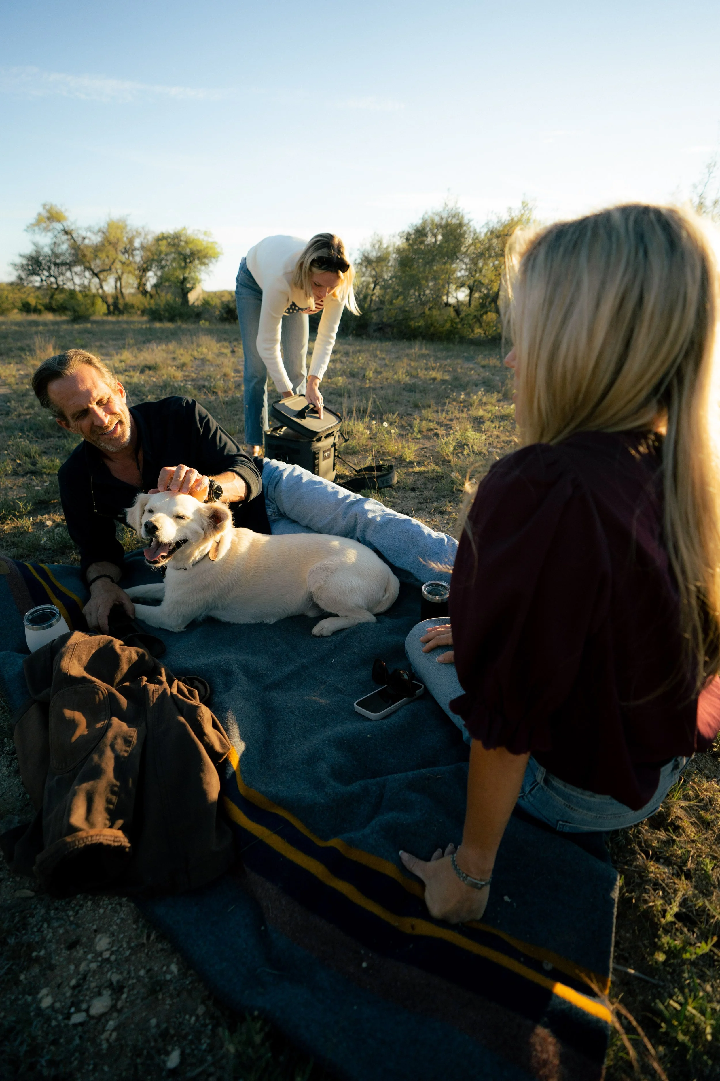 Four people and a dog outdoors in a grassy field during golden hour. One person is lying on the ground petting the dog, another is sitting nearby, and two others are standing and preparing for a picnic.