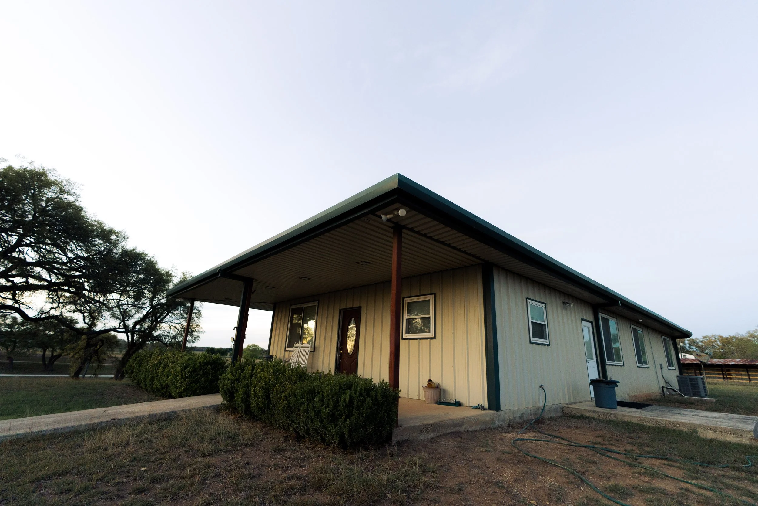 Rear view of a modern two-story house with a covered porch, a small set of stairs, trimmed bushes outside, and a tree on the left, under a cloudy sky.