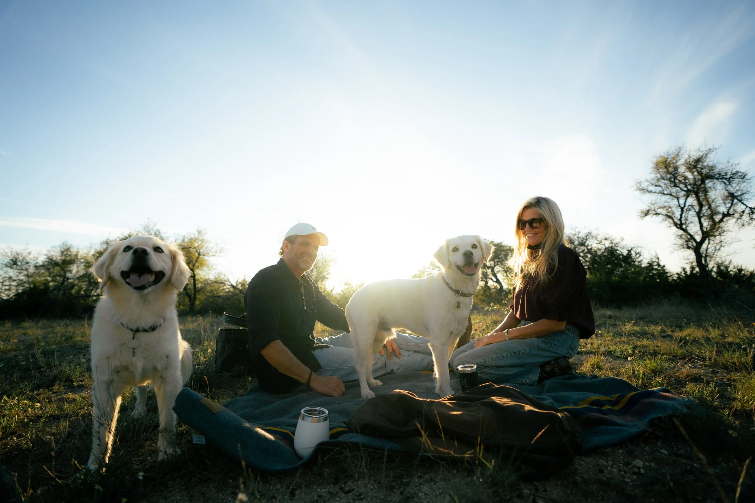 A man and woman sitting outdoors during sunset with two white dogs, one sitting and one standing, on a blanket in a grassy field with trees in the background.
