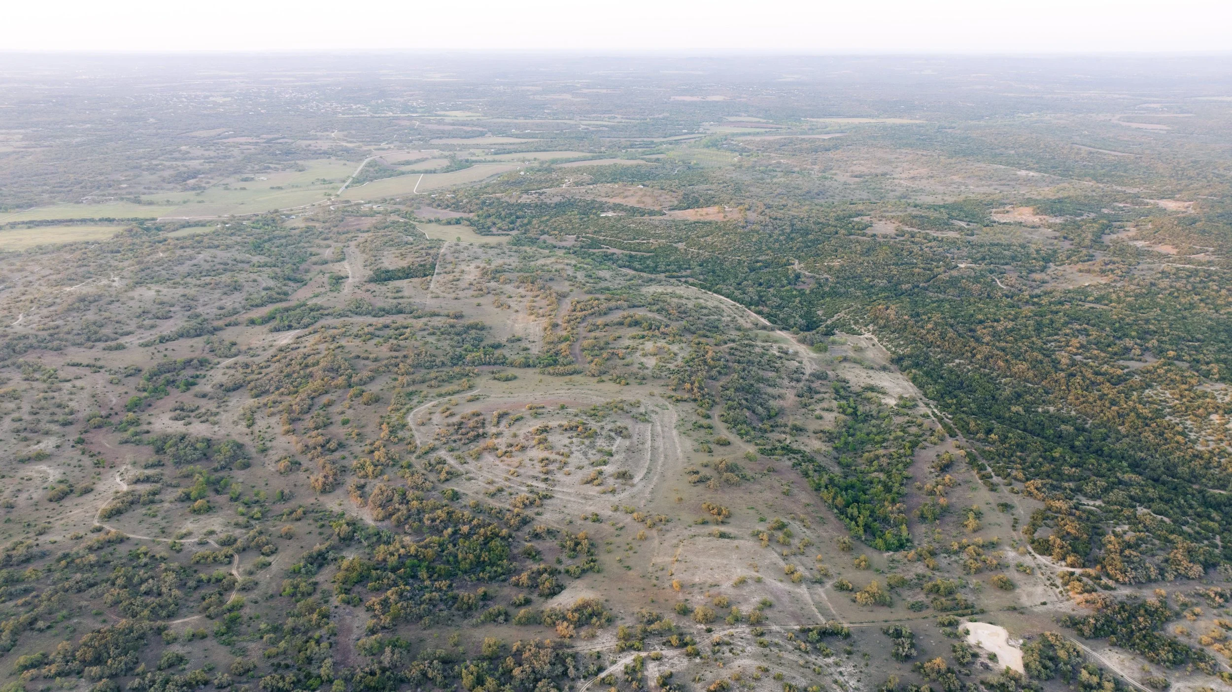 Aerial view of a landscape with fields, trees, and dirt roads stretching into the distance under a cloudy sky.