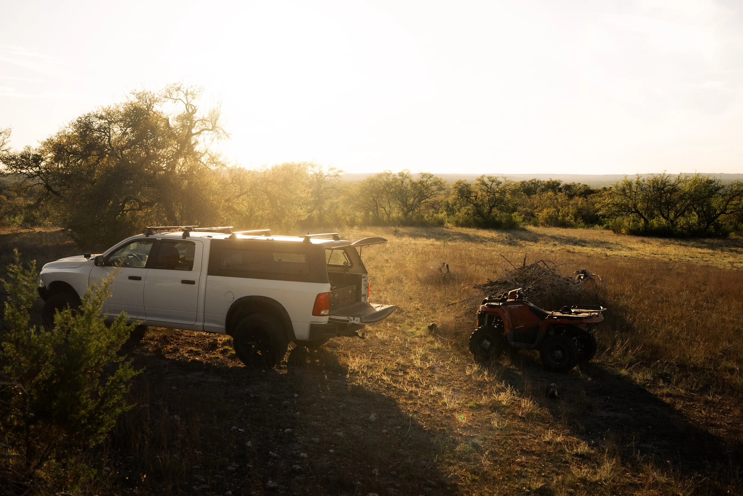A white pickup truck and a red all-terrain vehicle (ATV) parked on a grassy field with trees and bushes, backlit by the sun in a rural landscape.
