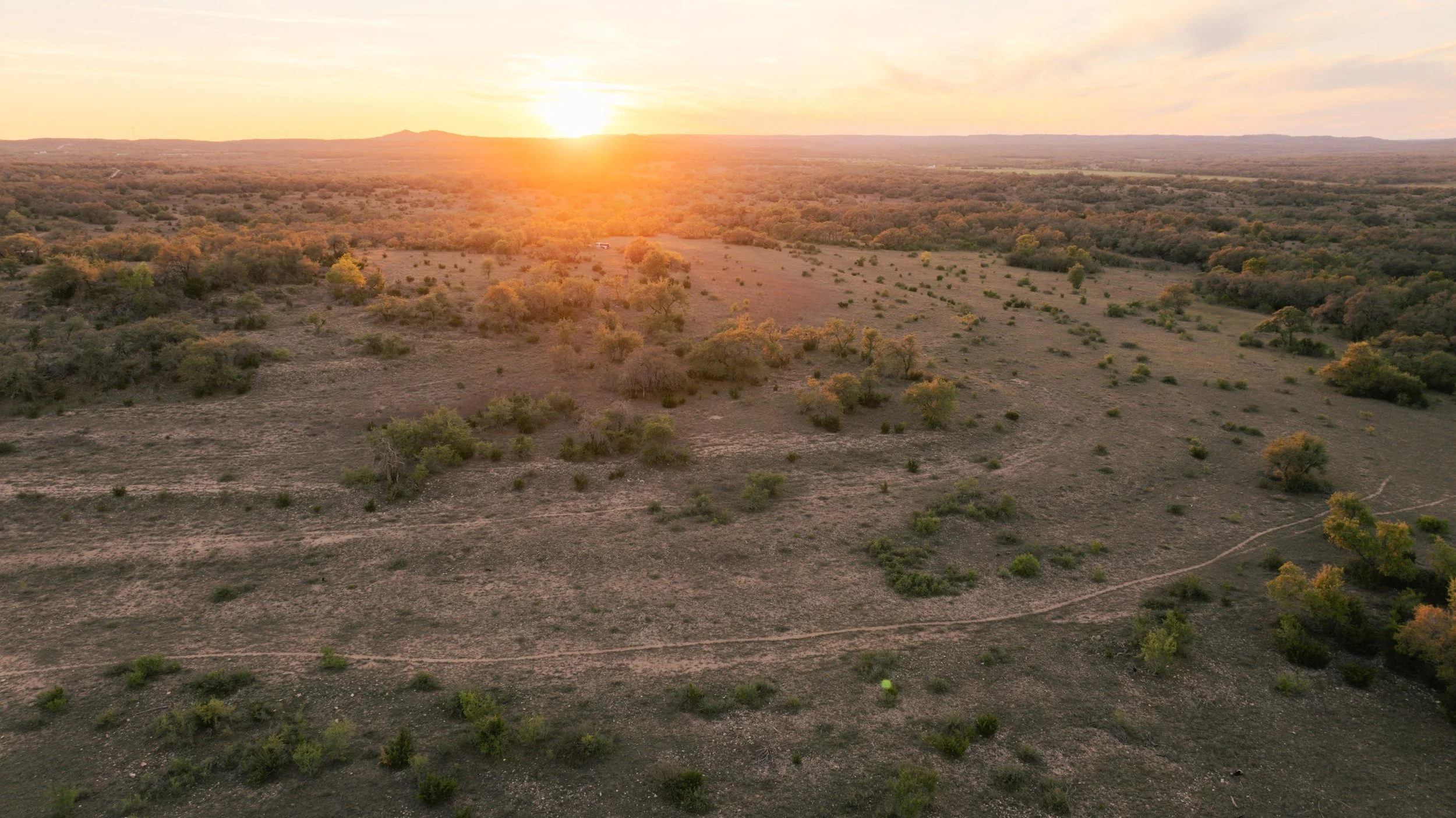 Sunset over a semi-arid landscape with sparse trees and bushes, rolling hills in the distance, and a dirt path in the foreground.
