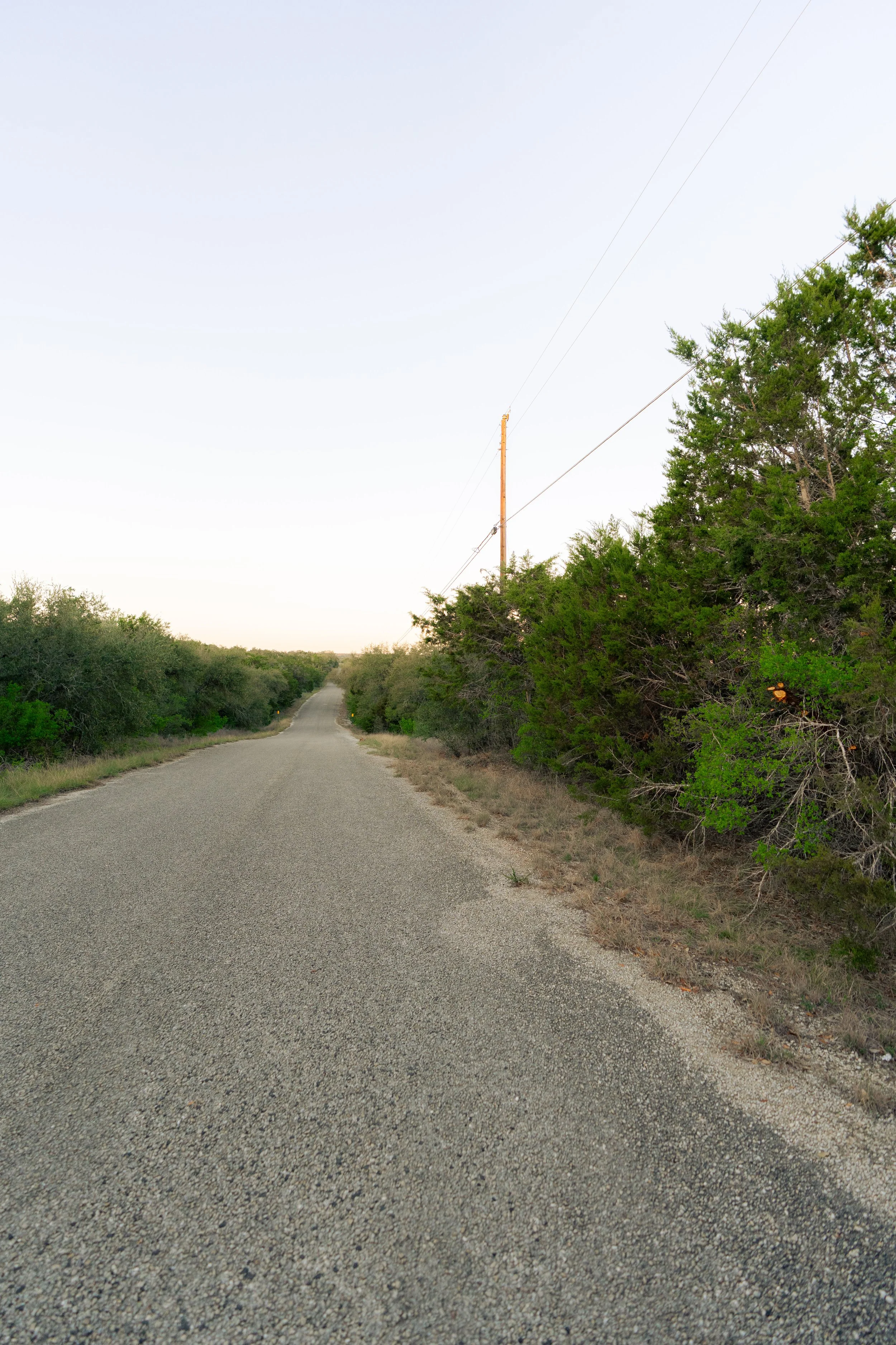 A long, straight gravel road running through a rural area with green bushes and trees on both sides and a utility pole with power lines on the right side.
