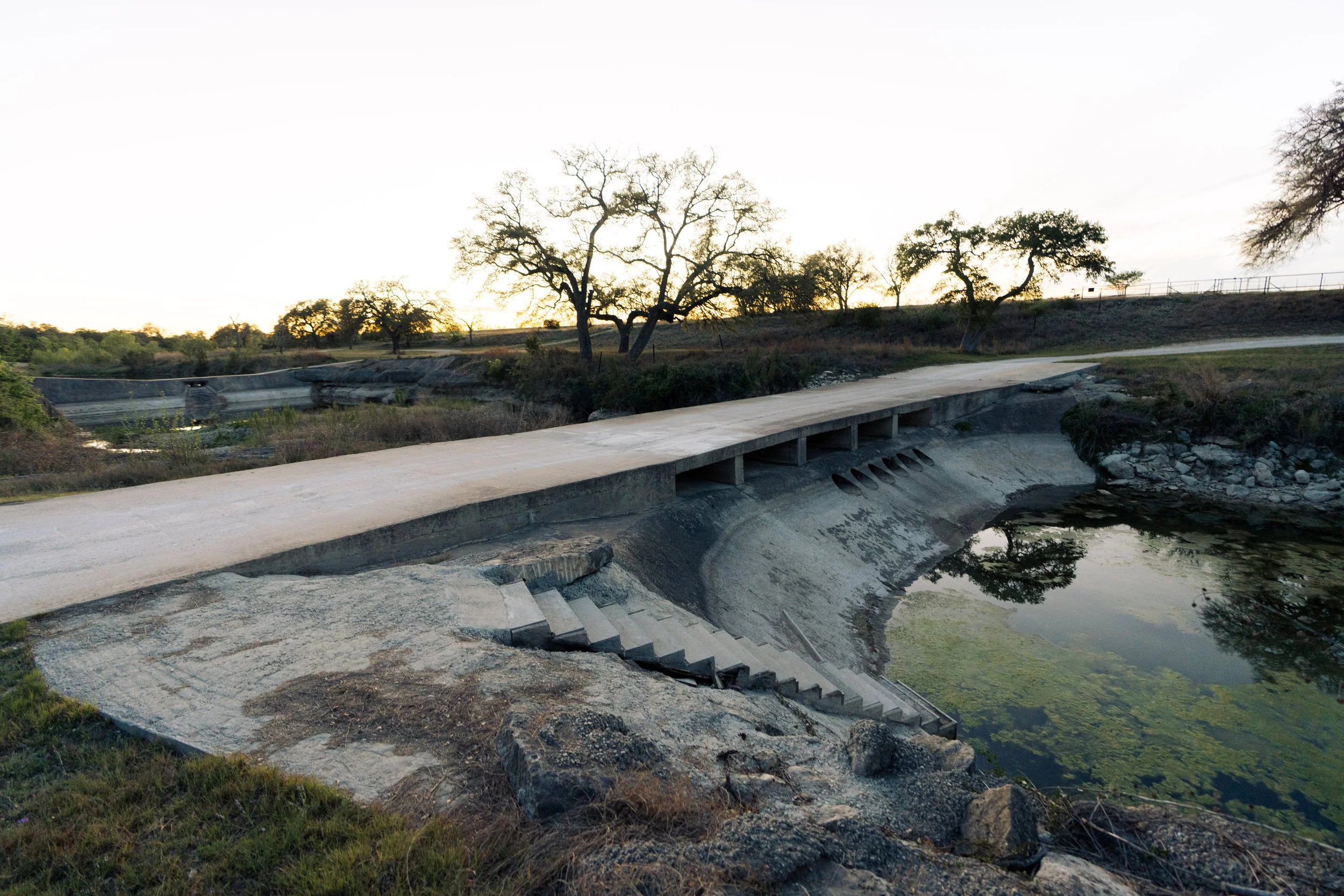 A broken concrete bridge over a waterway with trees in the background at sunset.