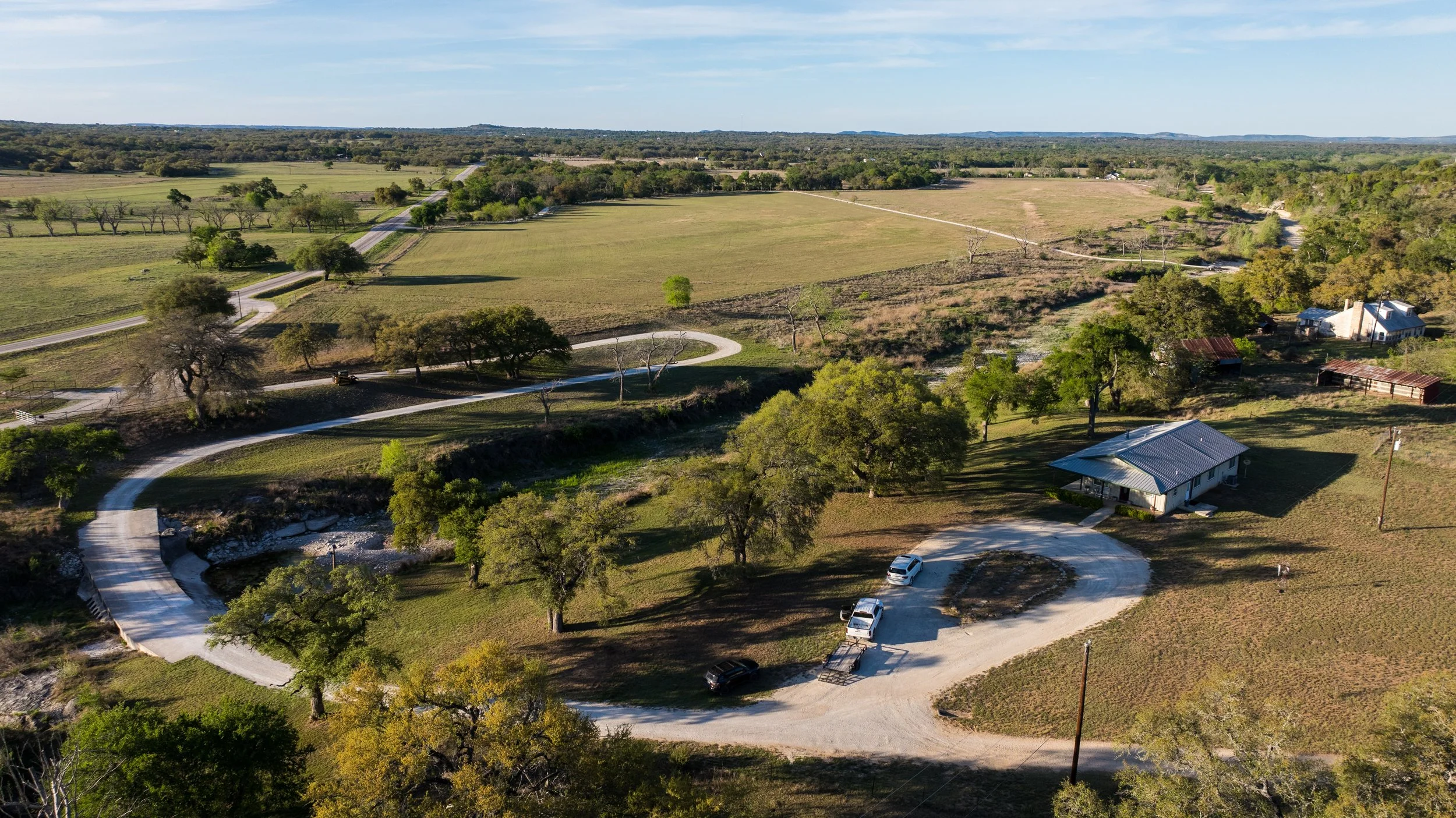 Aerial view of a rural farm with winding roads, houses, trees, open fields, and distant hills under a clear sky.