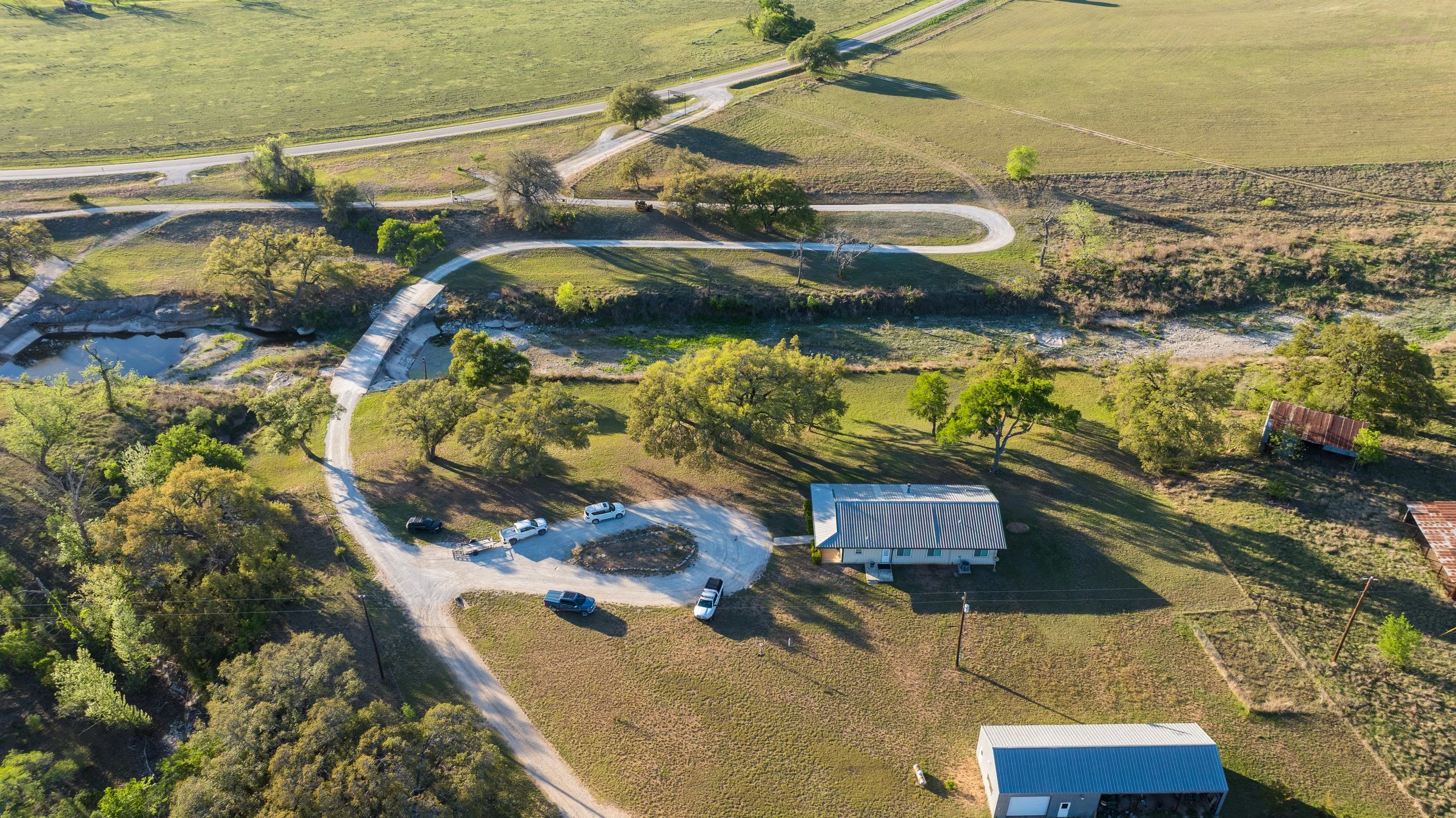 An aerial view of a rural property with a winding driveway, several parked cars, a small building with a metal roof, trees, and open fields.