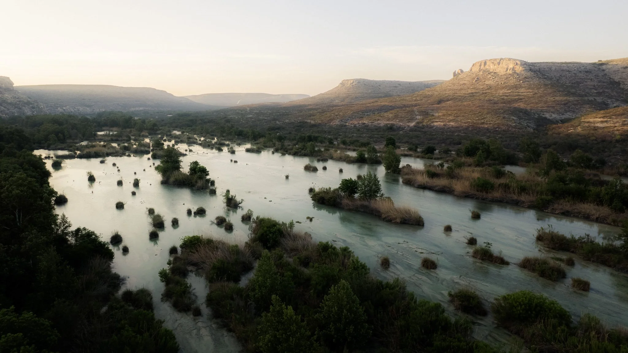 Landscape with a river flowing through green vegetation and bushes, surrounded by distant hills under a pale sky.