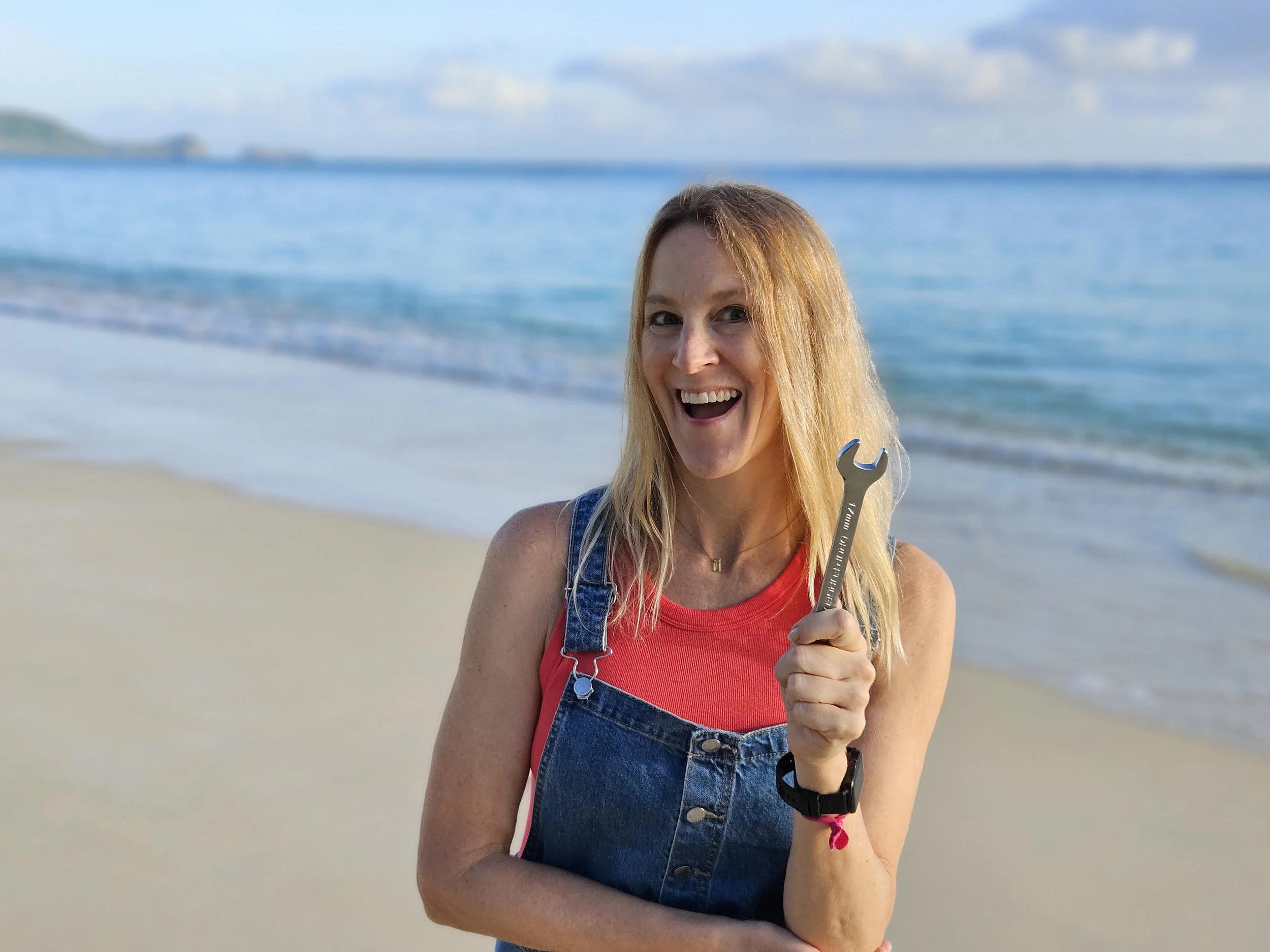 A woman in a red shirt and denim overalls smiling on a sandy beach near the ocean, holding a wrench, with sea and sky in the background.