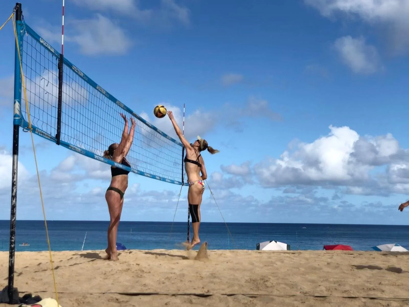 Women playing beach volleyball on sandy beach near ocean, under blue sky with scattered clouds, with umbrellas in the background.