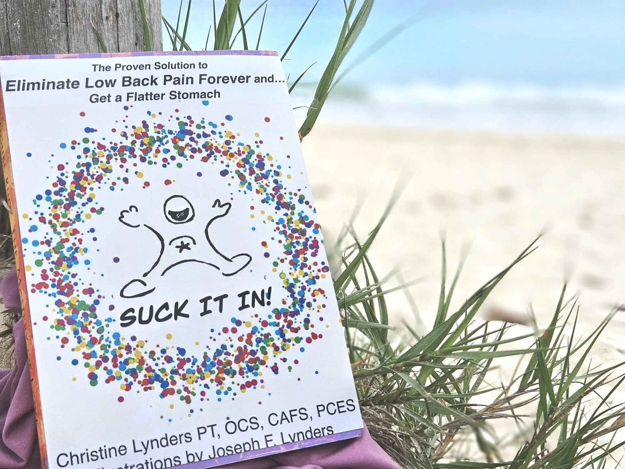A colorful flyer with a cartoon of a happy figure with arms raised, surrounded by multicolored confetti, and the words "SUCK IT IN!" at the bottom. The flyer advertises a solution for back pain and flatter stomachs, with partial text highlighting "Eliminate Low Back Pain Forever." It is placed amidst grass on a beach with sand and water in the background.