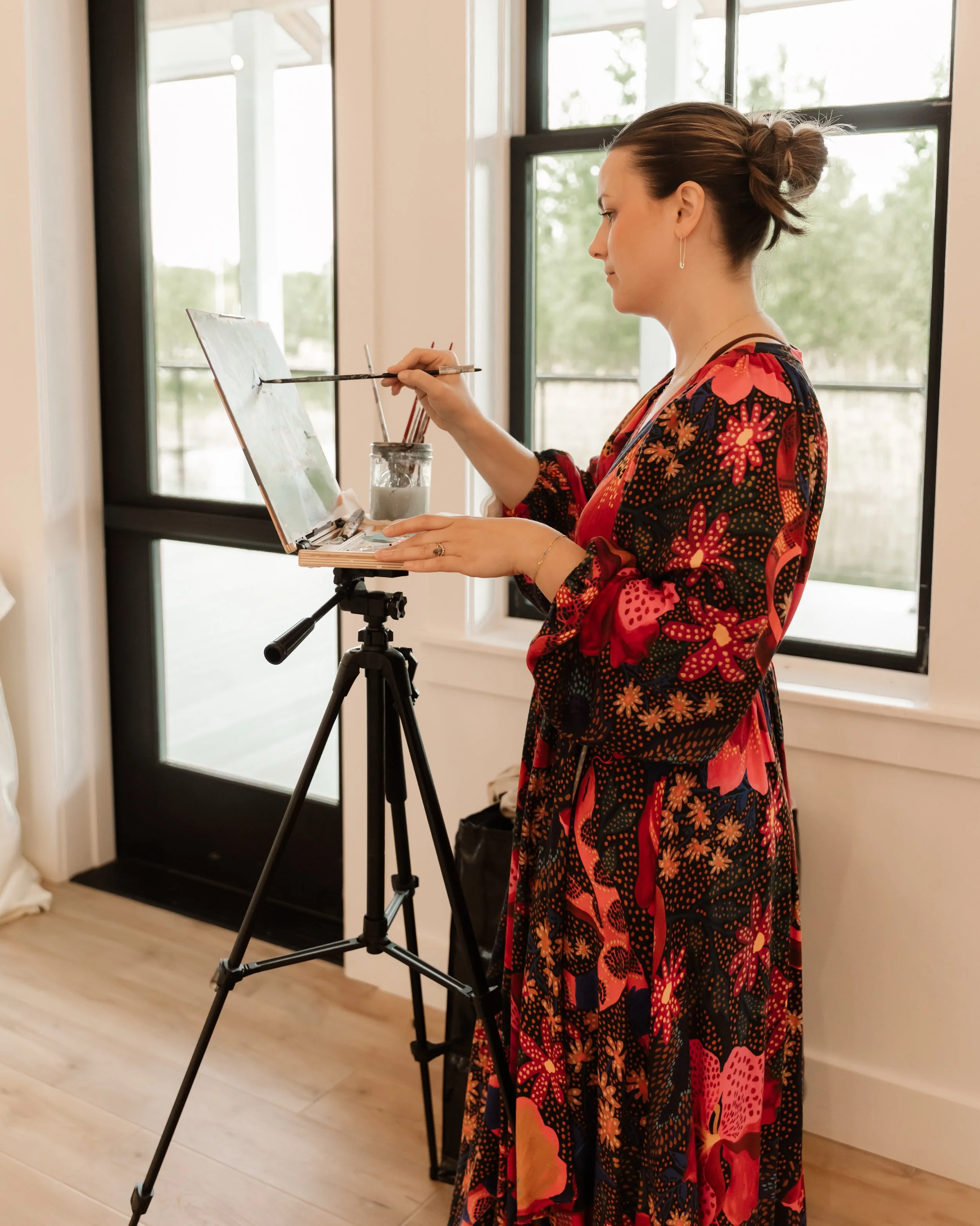 A female artist in a floral dress stands in front of a window, painting on a canvas set on a tripod easel, inside a bright room. new england art, coastal artist