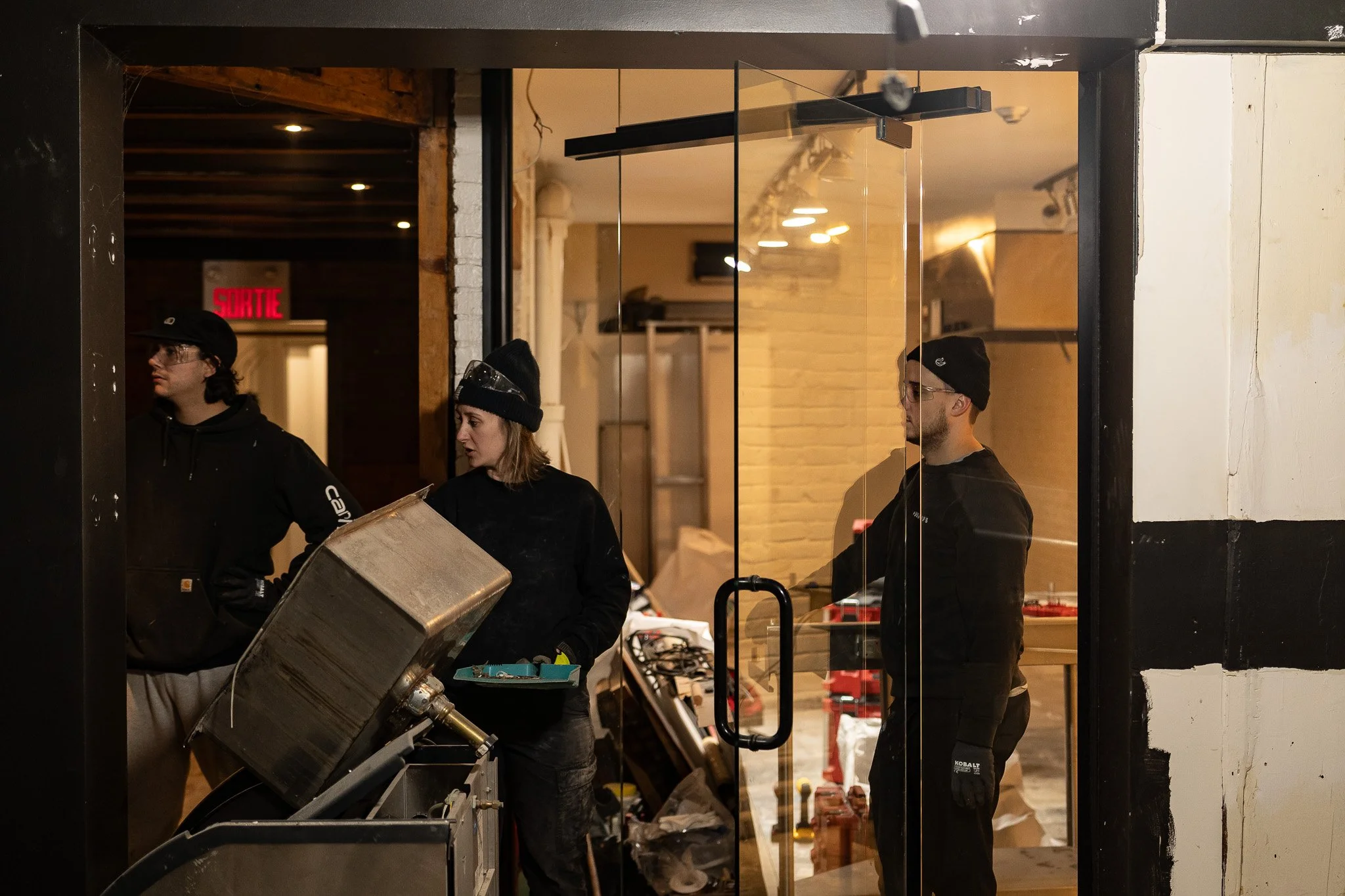 Trois personnes dans un atelier de construction ou rénovation, portant des vêtements de travail et des bonnets, près d'un équipement en métal.