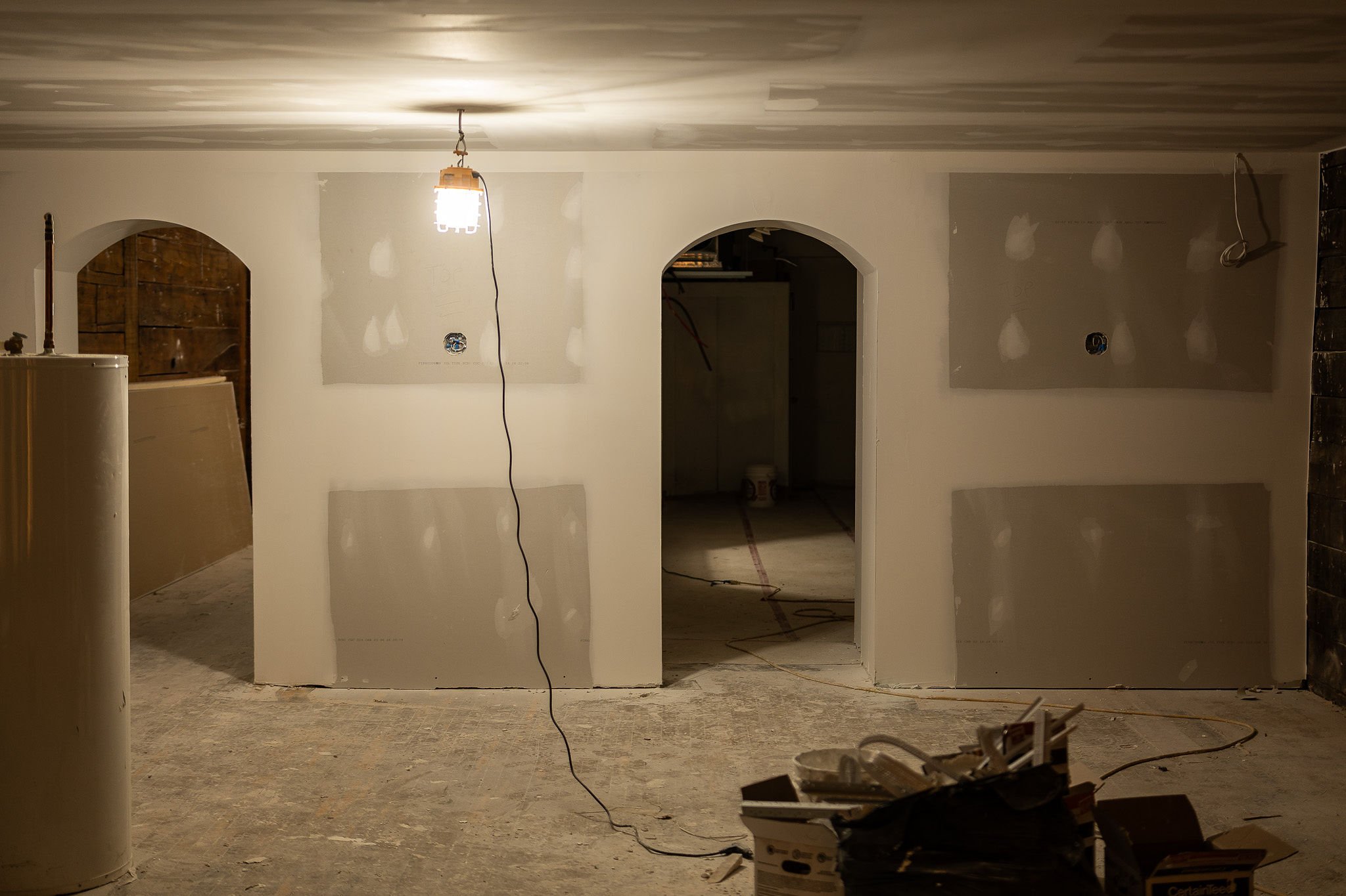 Salle en construction avec murs en placoplâtre, lumière suspendue, seau et matériel de construction au sol.