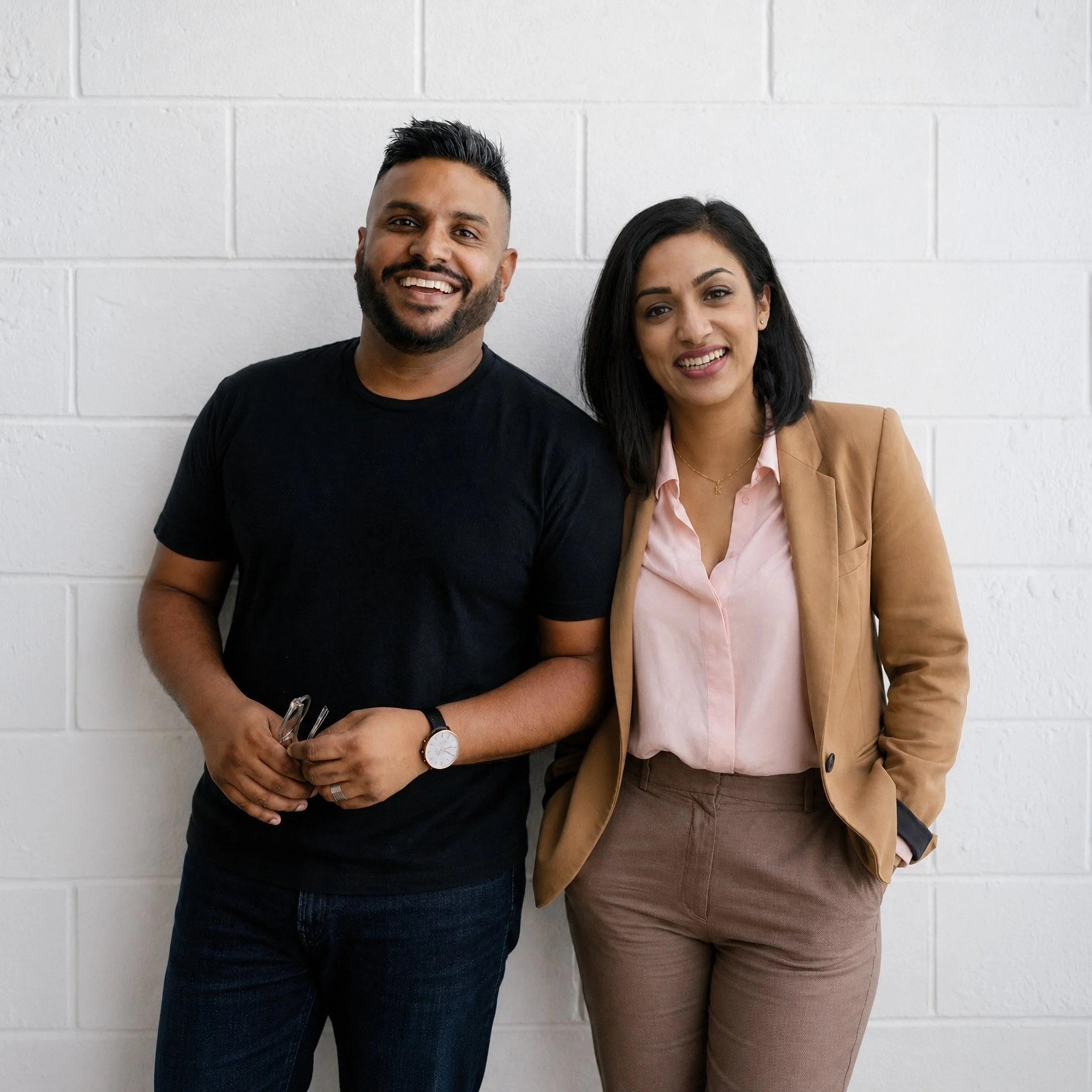 A smiling man and woman standing together against a white brick wall. The couple are Frank and Shruti Thiruchelvam, founders of Pink Tank Creative — a design and brand studio in Sydney, Australia.
