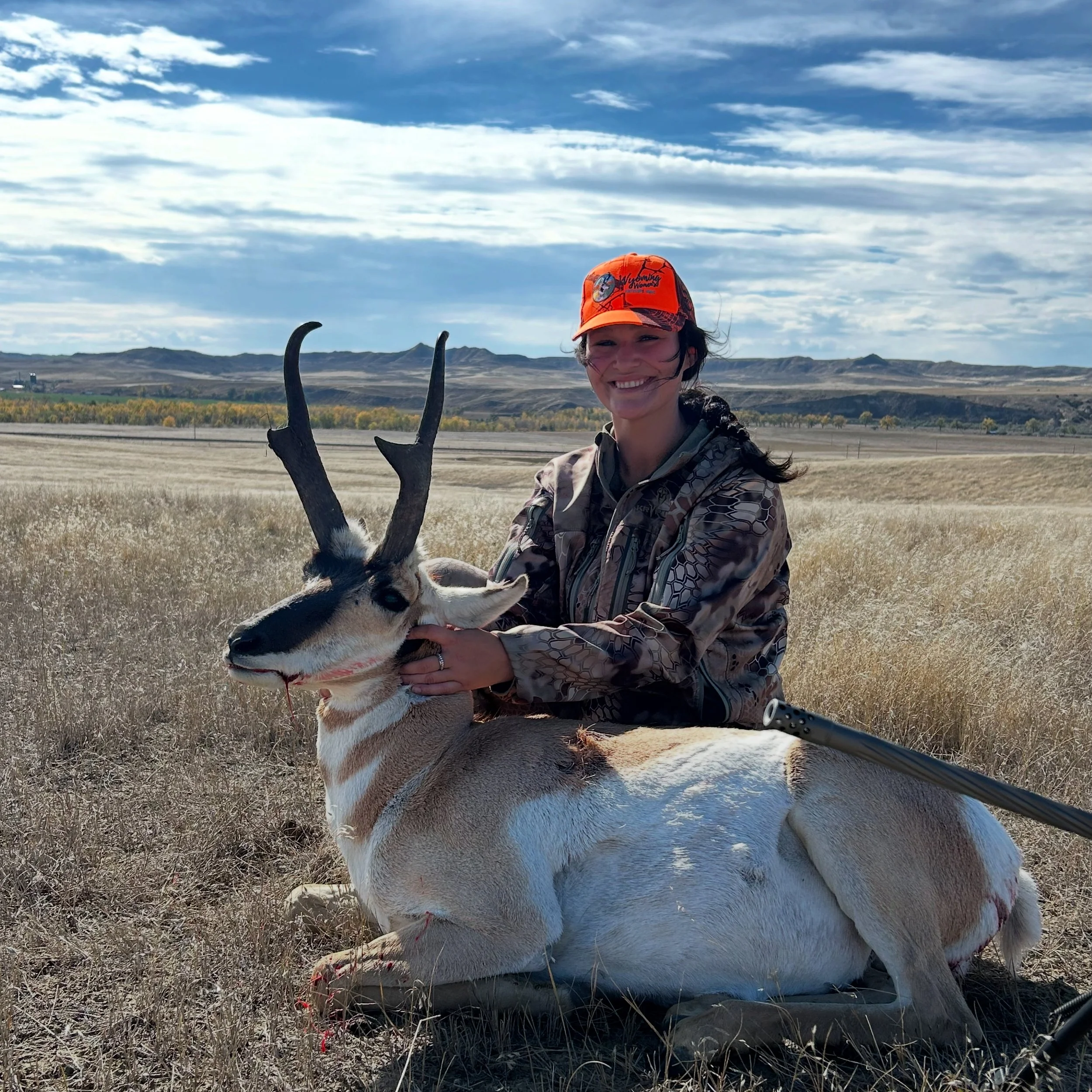 Woman in camouflage jacket and orange cap holding a pronghorn antelope in a field under a partly cloudy sky.