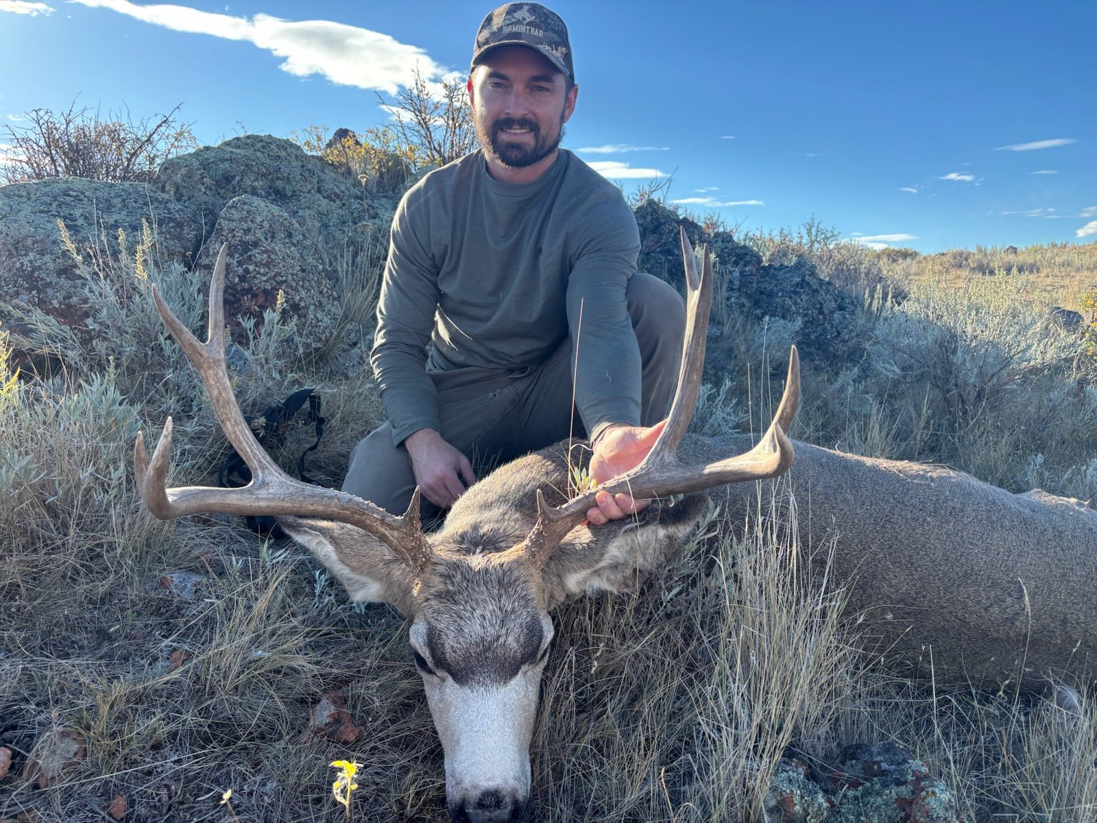A man in outdoor clothing and a cap squatting next to a large deer with antlers lying on the ground in a grassy area with rocks and bushes under a blue sky with clouds.