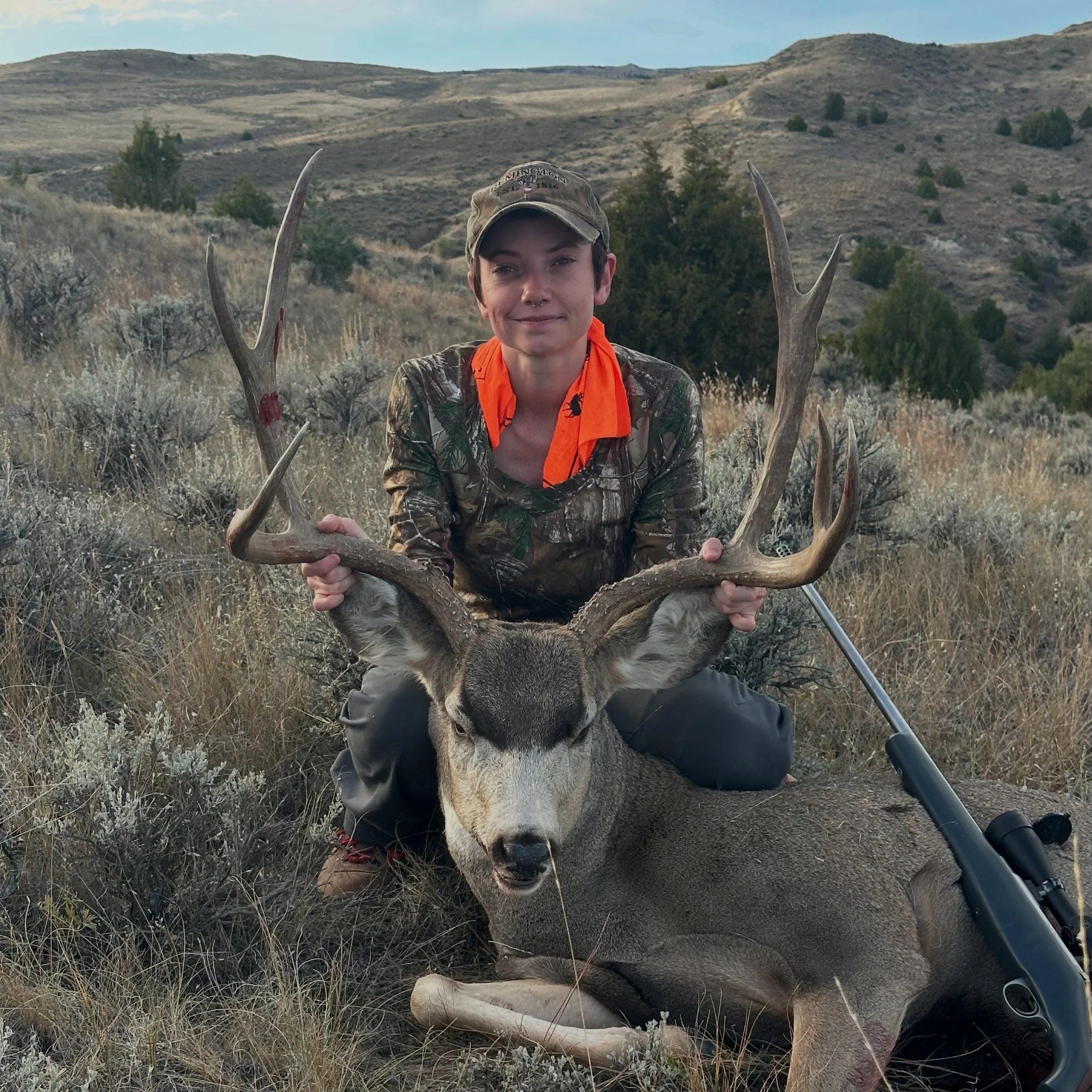 A person posing with a large deer that they have hunted, outdoors in a hilly, grassy landscape with bushes and trees.