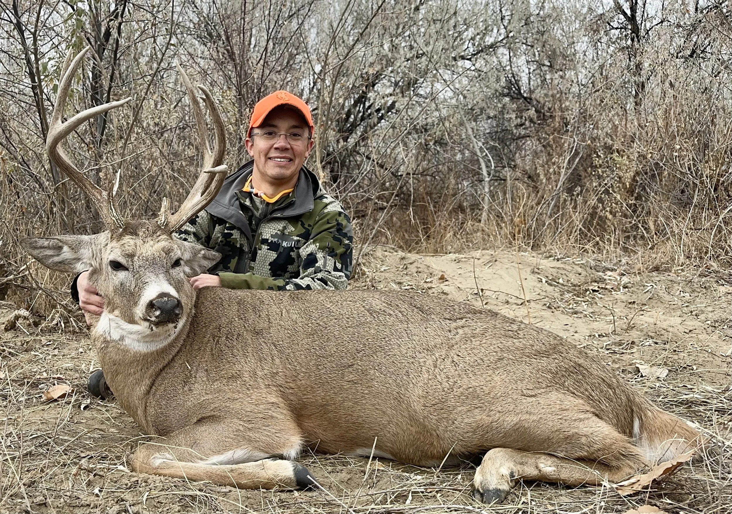 A man kneeling in dry brush holding a large dead deer with antlers in a forest setting.