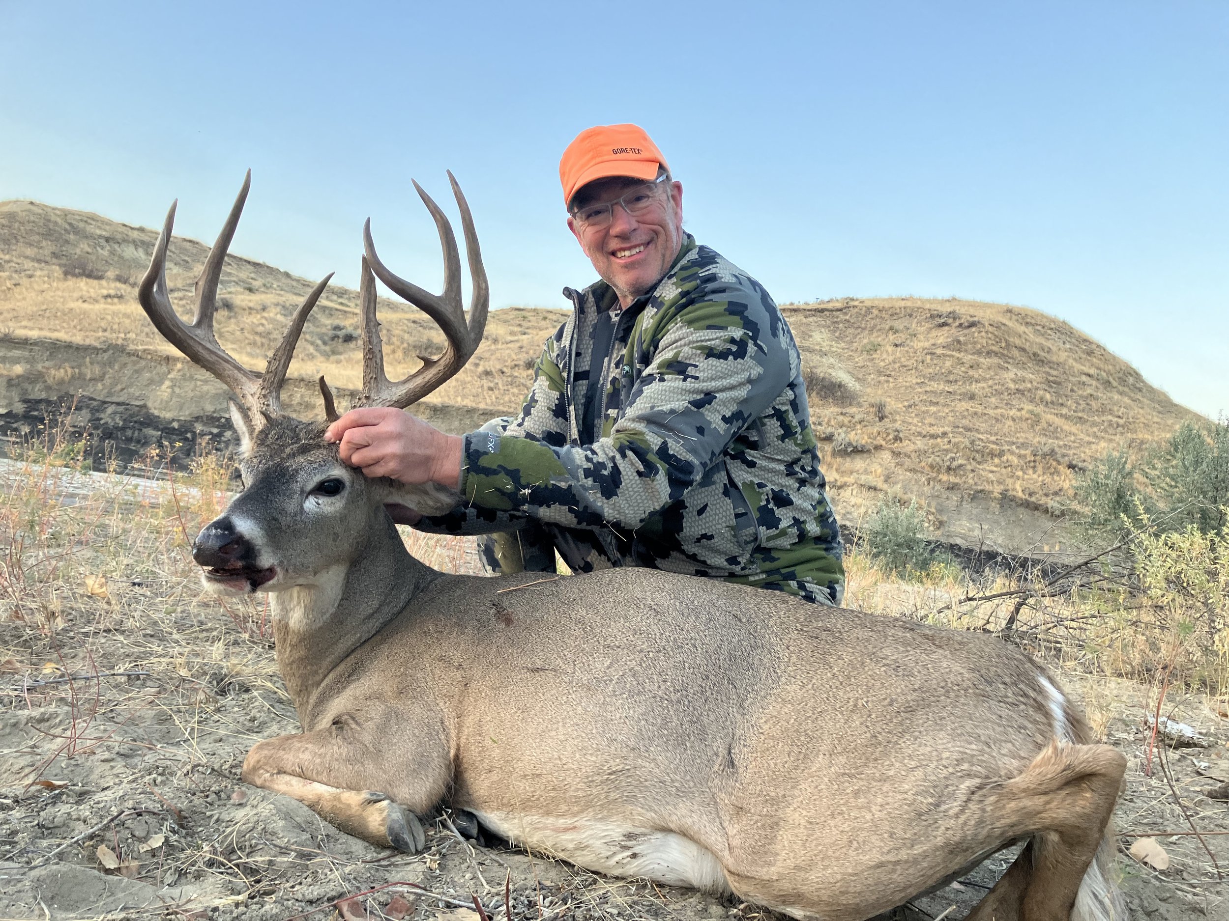 A man in camouflage clothing and an orange hat smiling while kneeling next to a large dead deer with impressive antlers, laying on dry ground with hills in the background.