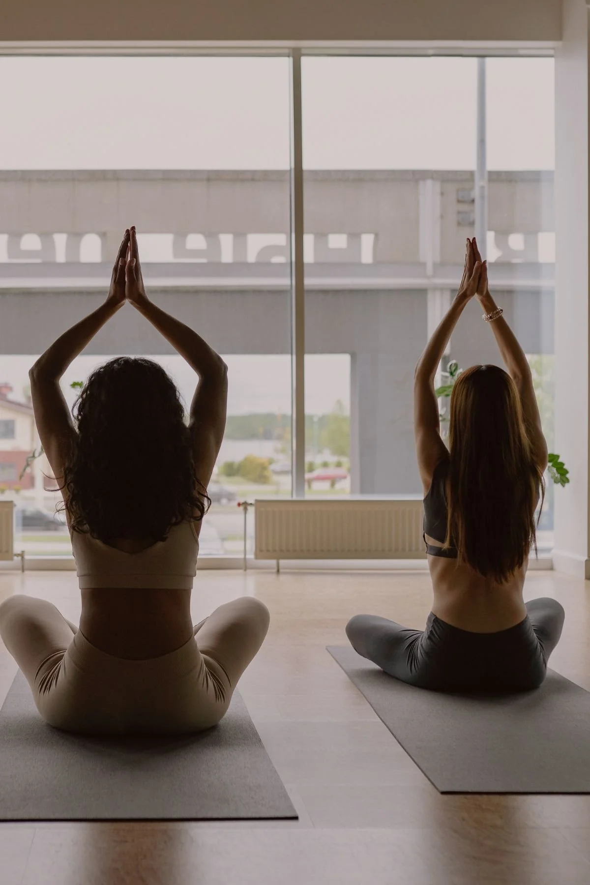 Two women practicing yoga indoors near a large window, sitting cross-legged with hands in prayer position above their heads.