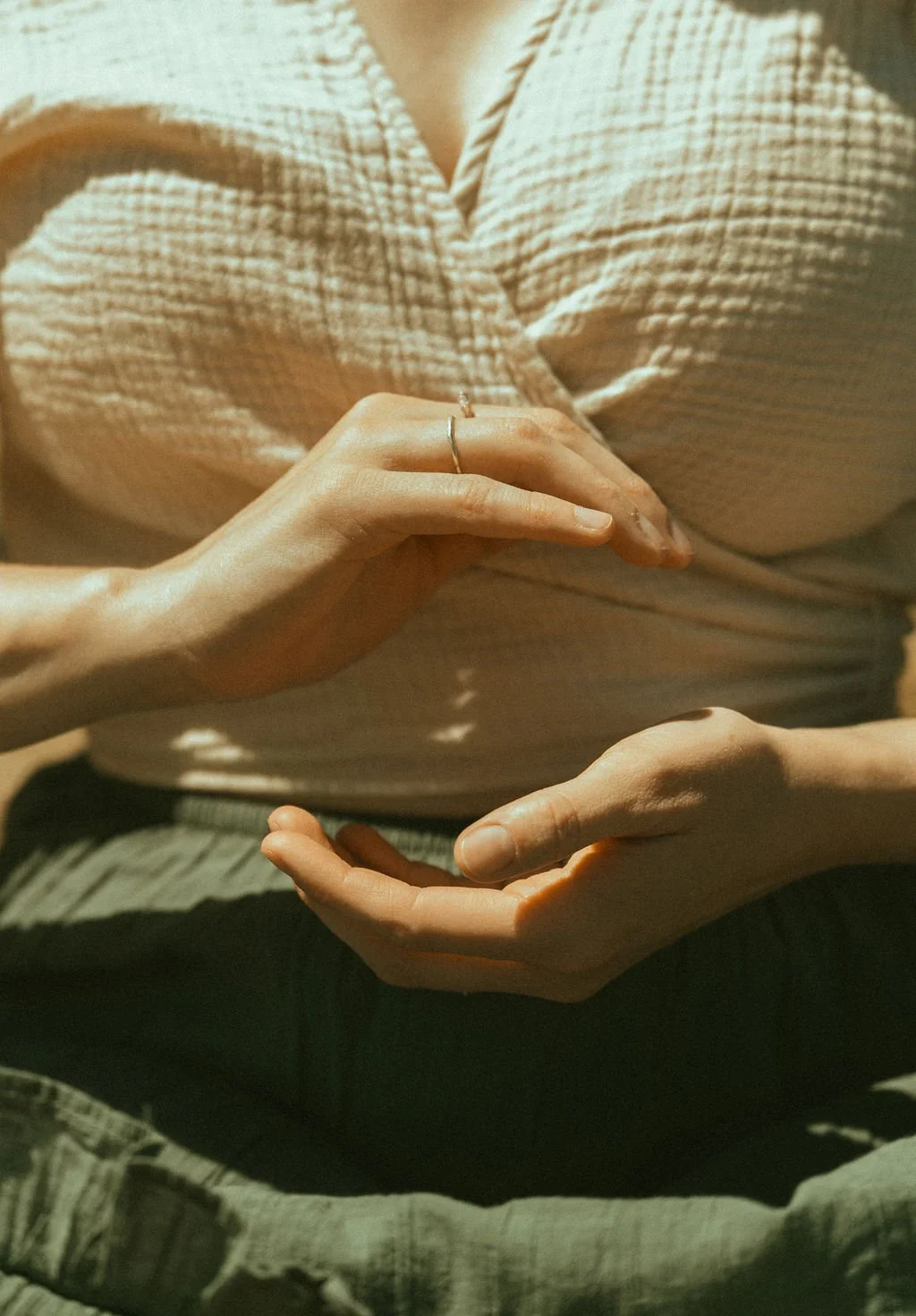 Close-up of a person's hands, one hand resting on their lap and the other hand placed on their stomach. The person is wearing a beige, textured top and olive green pants.