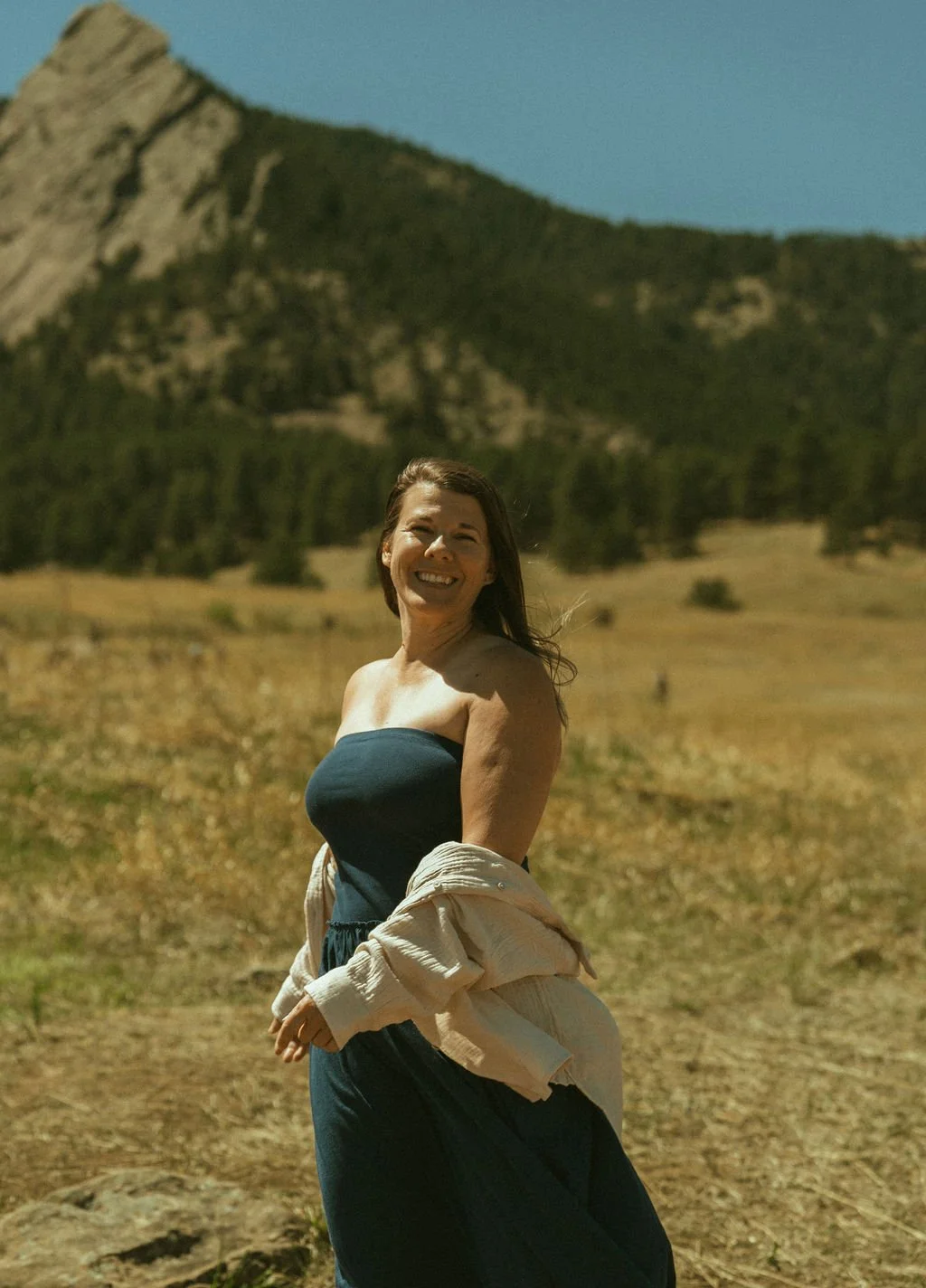 A smiling woman standing outdoors in a mountain landscape with dry grass and trees under a blue sky.