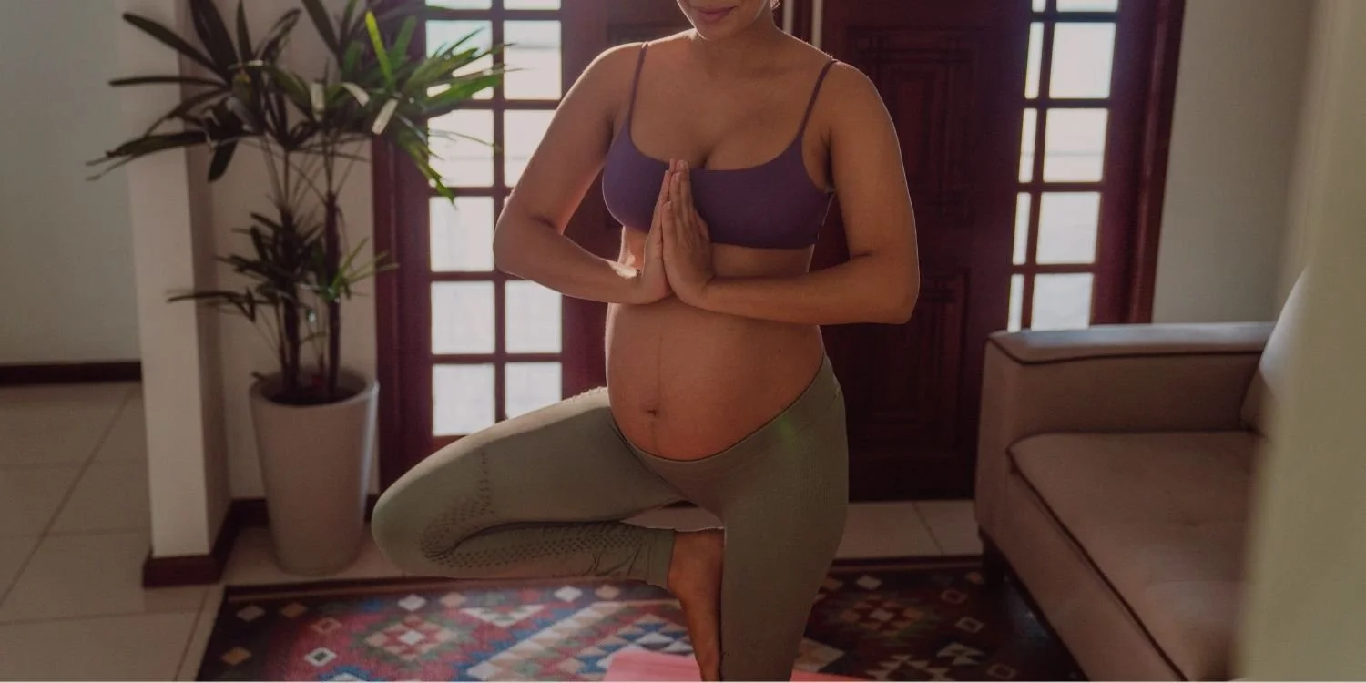 A pregnant woman practicing yoga indoors, standing on one leg in a tree pose, with hands in prayer position, wearing a purple sports bra and beige leggings.