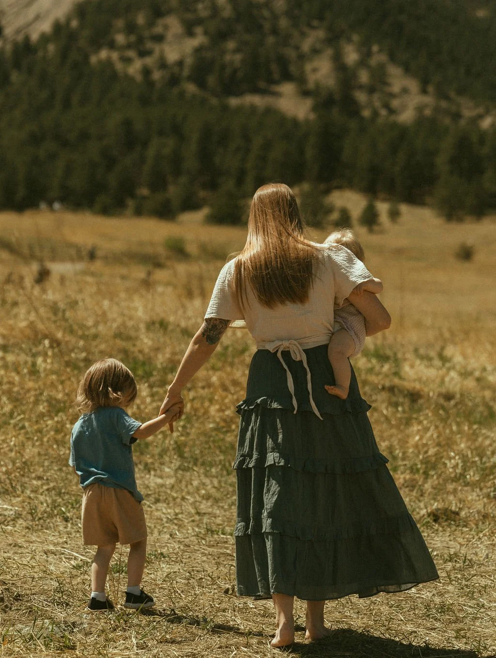 A woman walking with two young children in a dry, grassy field with mountains in the background. One child is holding the woman's hand, and the other is being carried by her.