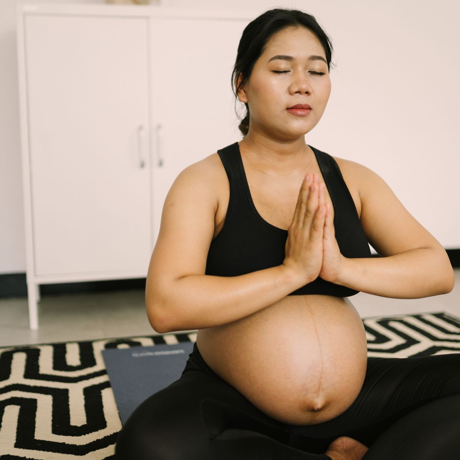 Pregnant woman practicing yoga in a black sports bra and black pants, seated on a patterned black and white rug with eyes closed and hands in a prayer position.