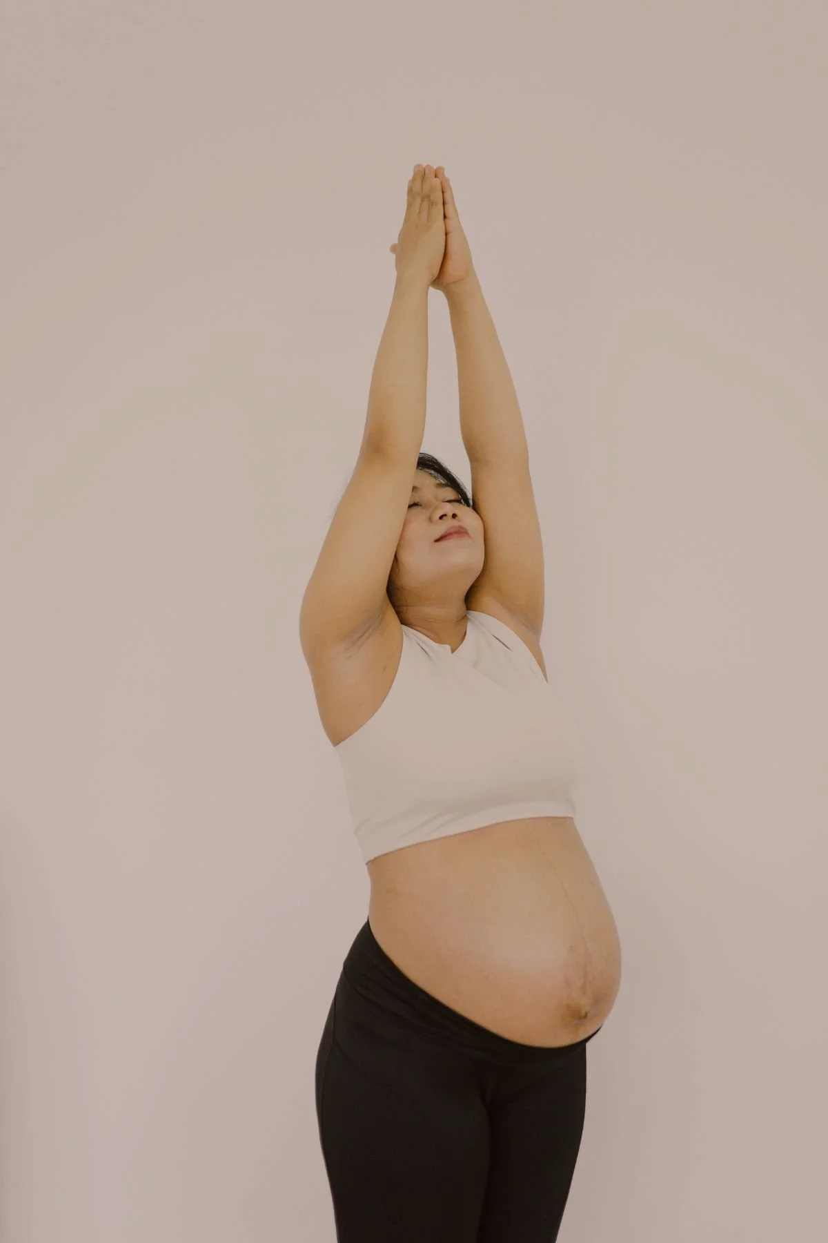 A woman practicing yoga with her arms extended upward and hands pressed together in a prayer position, standing against a plain light-colored wall.