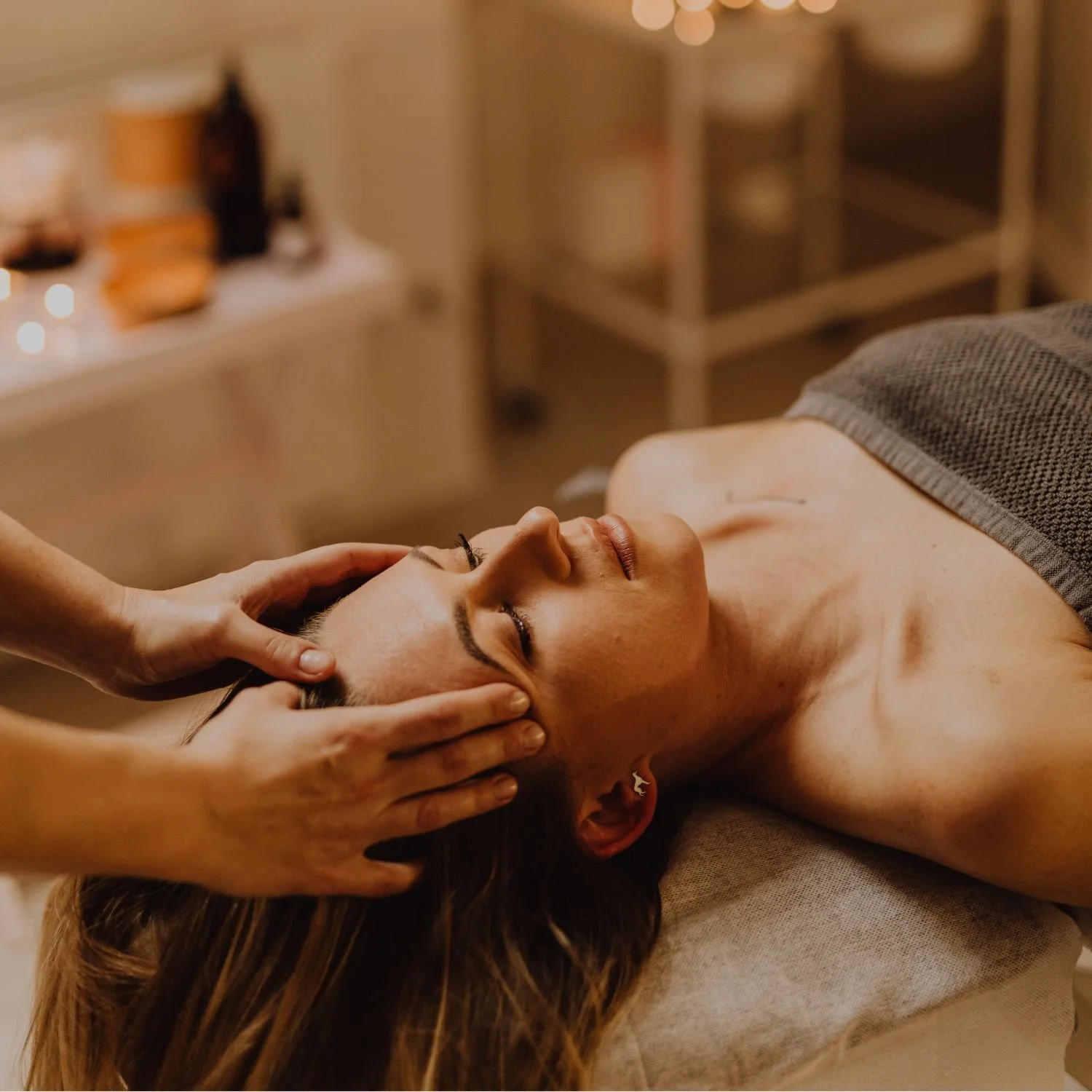 A woman receives a facial massage at a spa, lying on a massage table with her eyes closed and a towel covering her chest, in a dimly lit room.